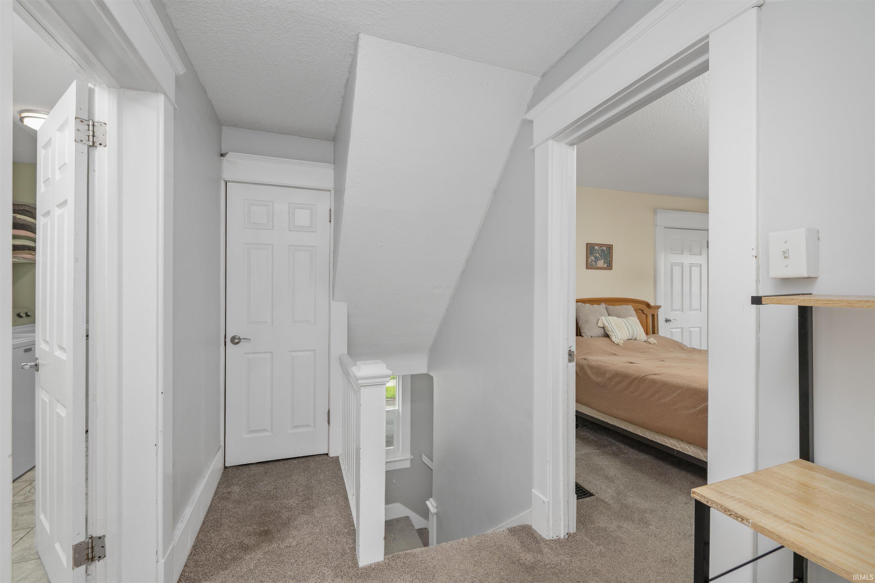 Carpeted hallway featuring white trim and panel doors, with a visible bedroom entrance and a partially visible laundry area