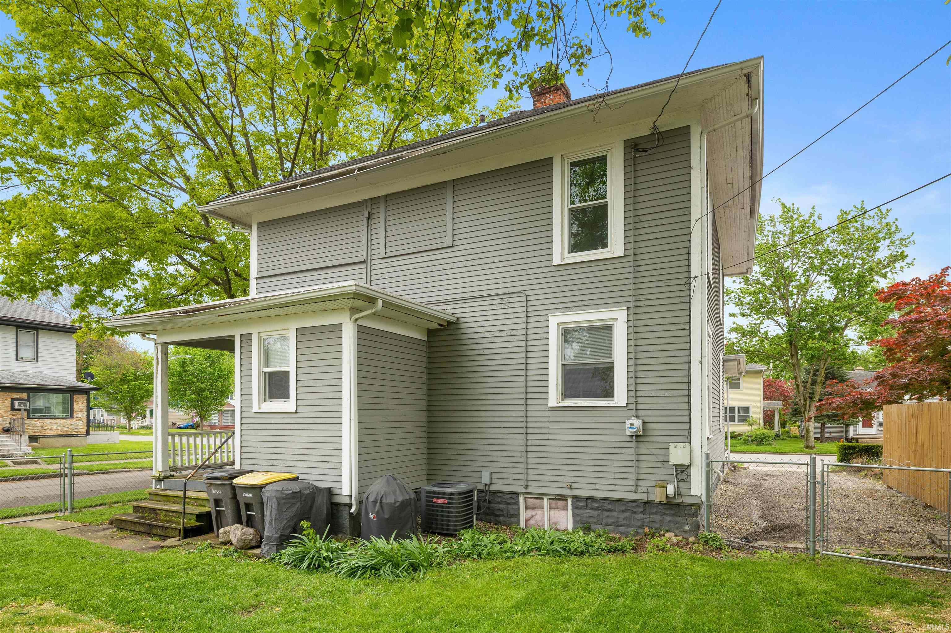 Two-story exterior featuring gray siding, a covered porch, a manicured lawn, and a fenced gravel driveway