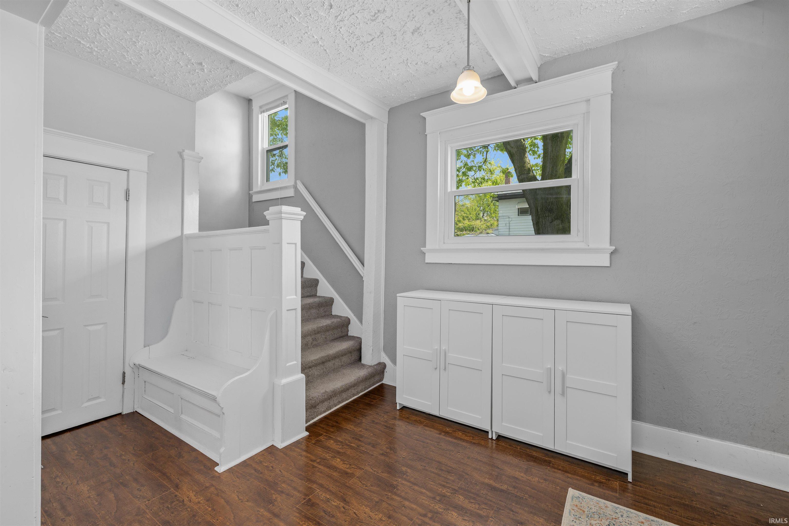 Entryway featuring dark wood-finish flooring, a built-in white bench, a stairway with carpeted treads, a window with white trim, and white storage cabinets