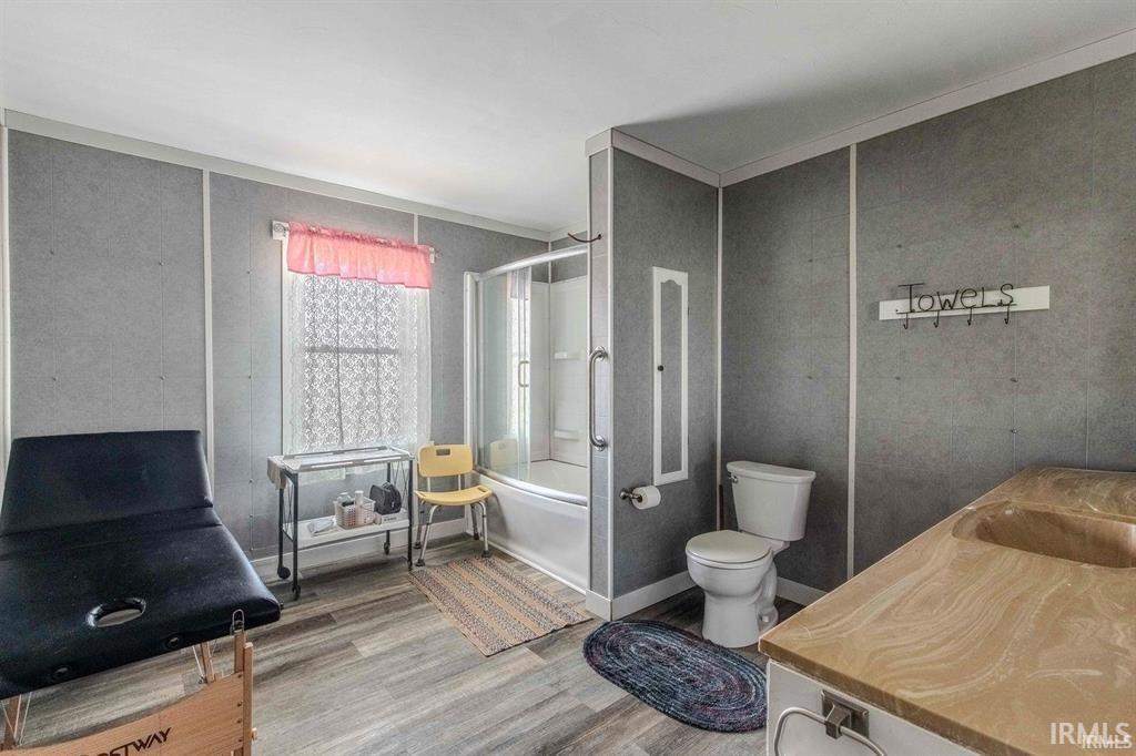 Bathroom featuring wood-finish flooring, a combination tub and shower with glass enclosure, a vanity with an integrated sink, a toilet, and a window with privacy covering