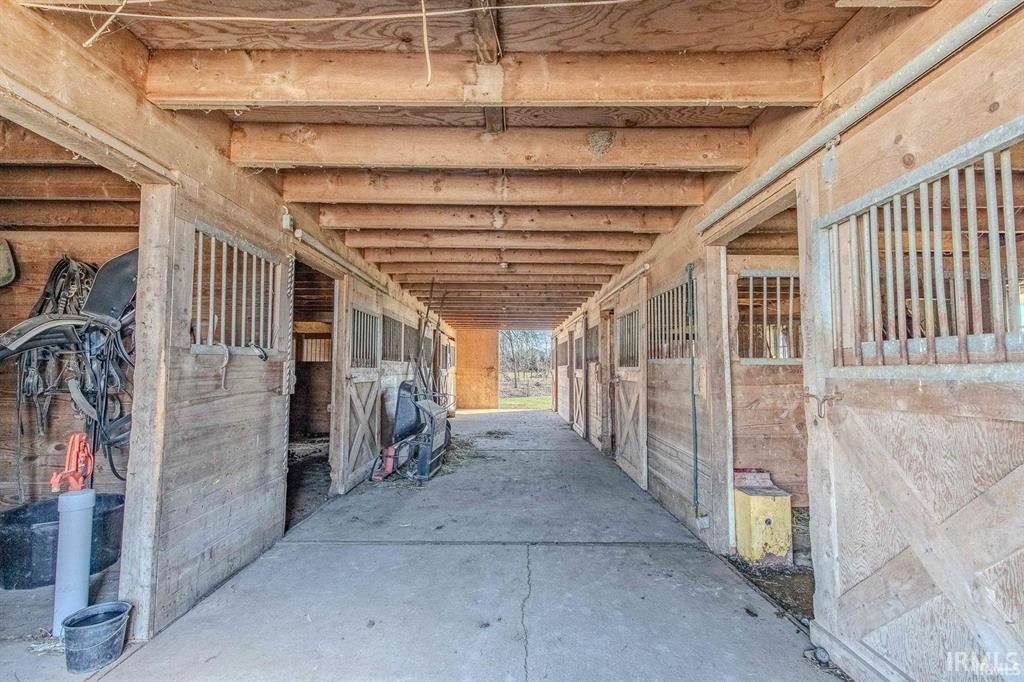 Barn interior featuring multiple stalls with sliding wood doors and metal grates, exposed wood beam ceiling, and a concrete aisle
