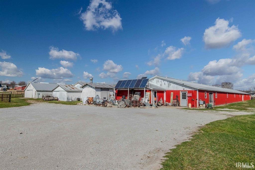 Expansive gravel parking area fronting multiple outbuildings, including a prominent red-sided structure with solar panels and a white entrance door