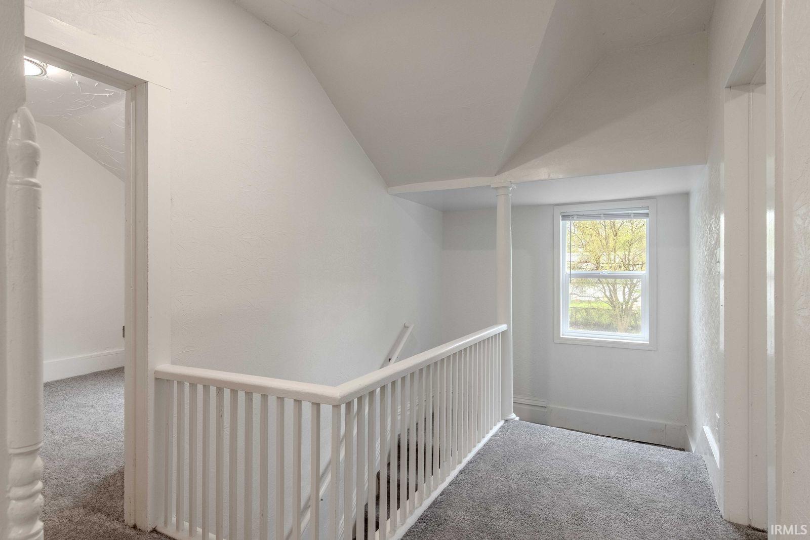 Carpeted hallway featuring a white spindle railing, a single window with white trim, and a white interior column