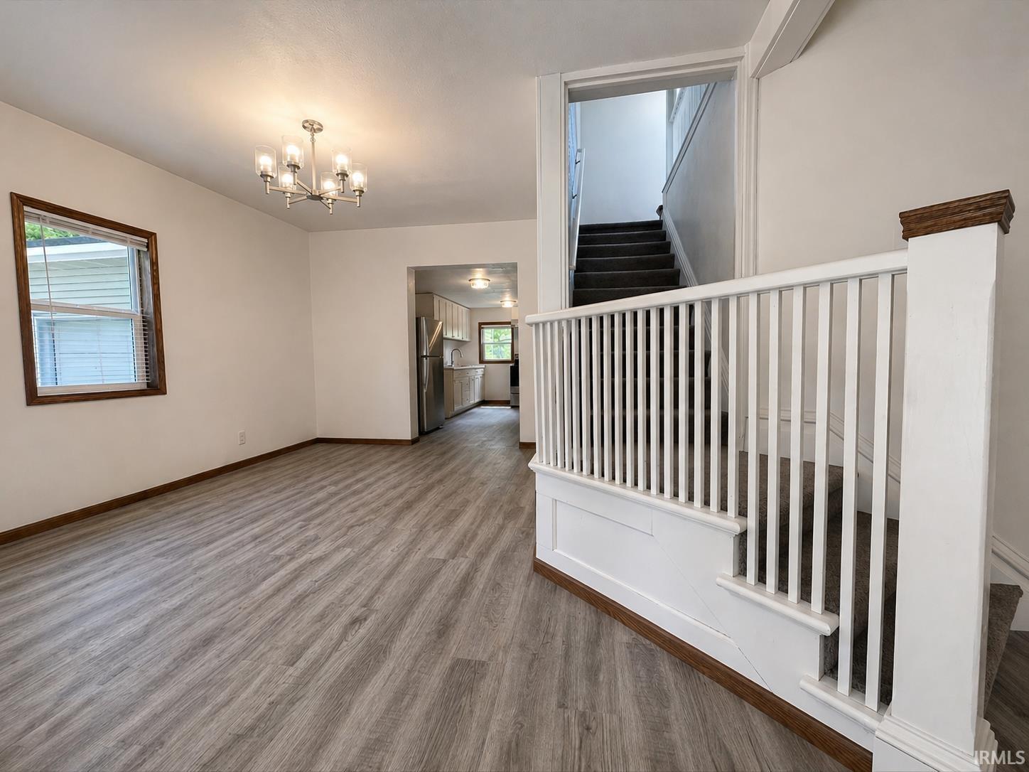 Spacious living area featuring wood-finish flooring, a modern chandelier, and ample natural light from a window with wood trim