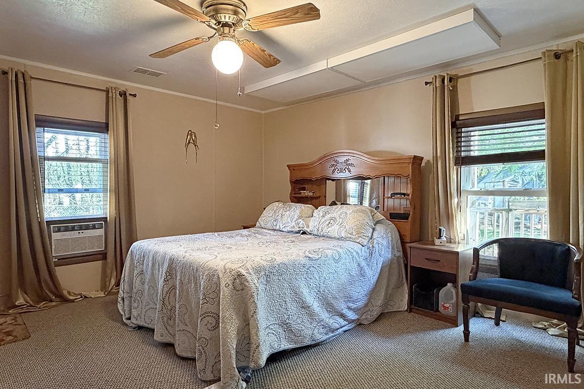Carpeted bedroom featuring two large windows with blinds and drapes, a ceiling fan with integrated lighting, and crown molding