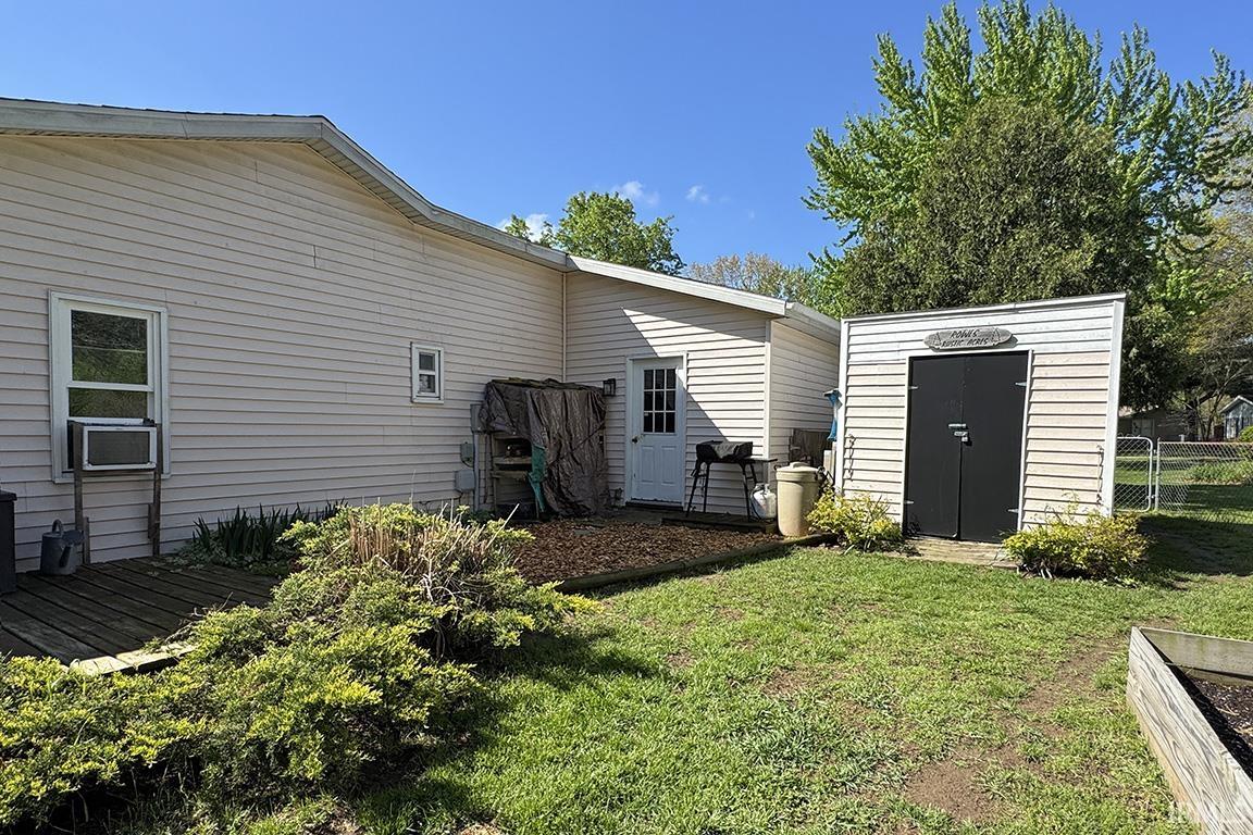 Rear exterior featuring a white vinyl siding, an outdoor storage shed with black doors, a wooden deck, a secondary structure with a white entry door, and a raised garden bed