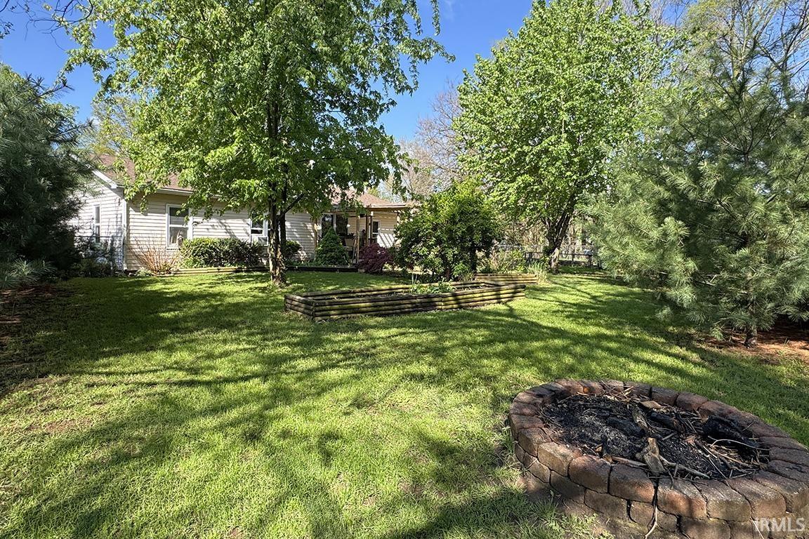 Expansive grassy yard featuring a brick-built fire pit, mature trees, and a raised garden bed