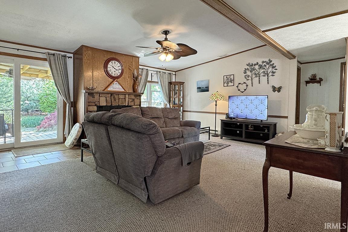 Living area featuring a stone-faced fireplace with wood paneling, sliding glass doors to outdoor space, ceiling fan, carpet flooring, and exposed ceiling beams