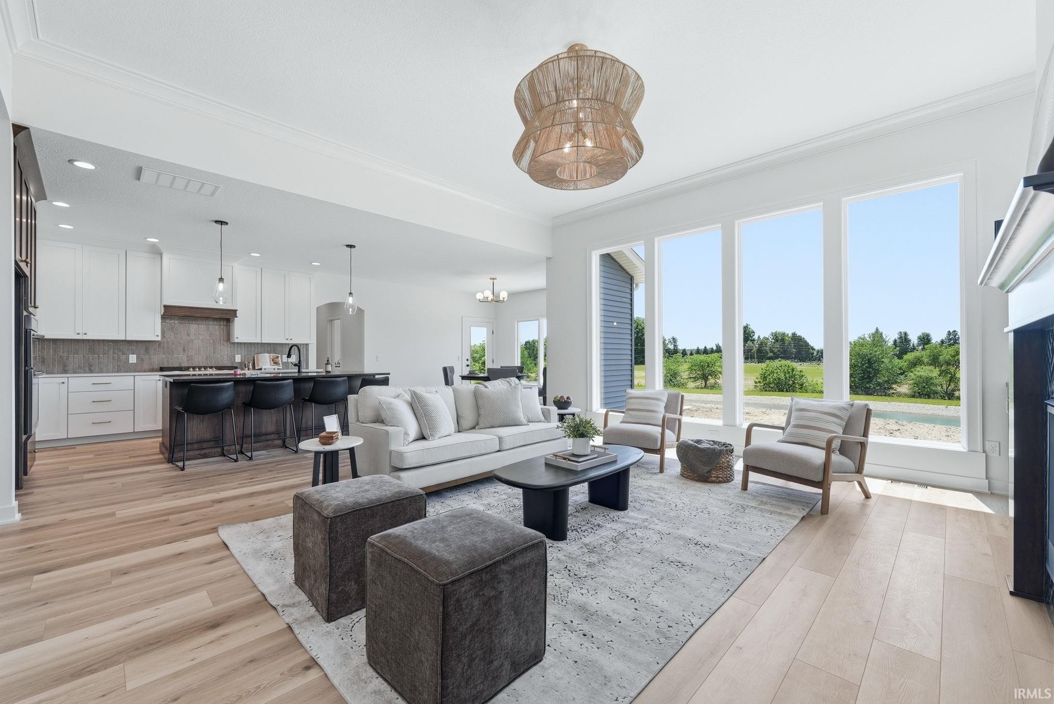 Open-concept living space featuring wood-finish flooring, a contemporary kitchen with white cabinetry and a dark island, crown molding, a decorative ceiling fixture, and a wall of tall windows