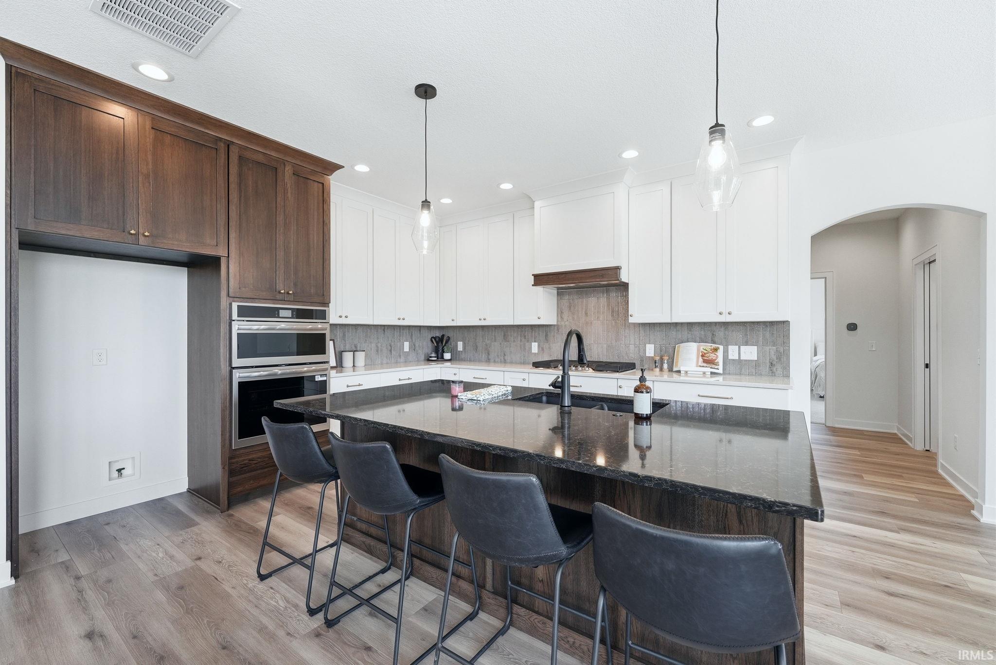 Modern kitchen featuring an expansive island with a dark countertop, white upper cabinetry, rich wood-finish lower cabinetry and built-ins, stainless steel wall oven and microwave, and wood-finish flooring