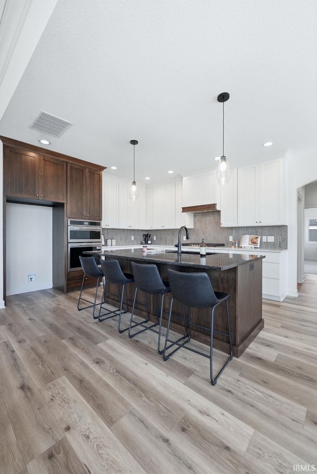 Gourmet kitchen featuring a large island with dark stone countertop, white upper cabinetry, dark wood lower cabinetry, stainless steel double ovens, and wood-finish flooring