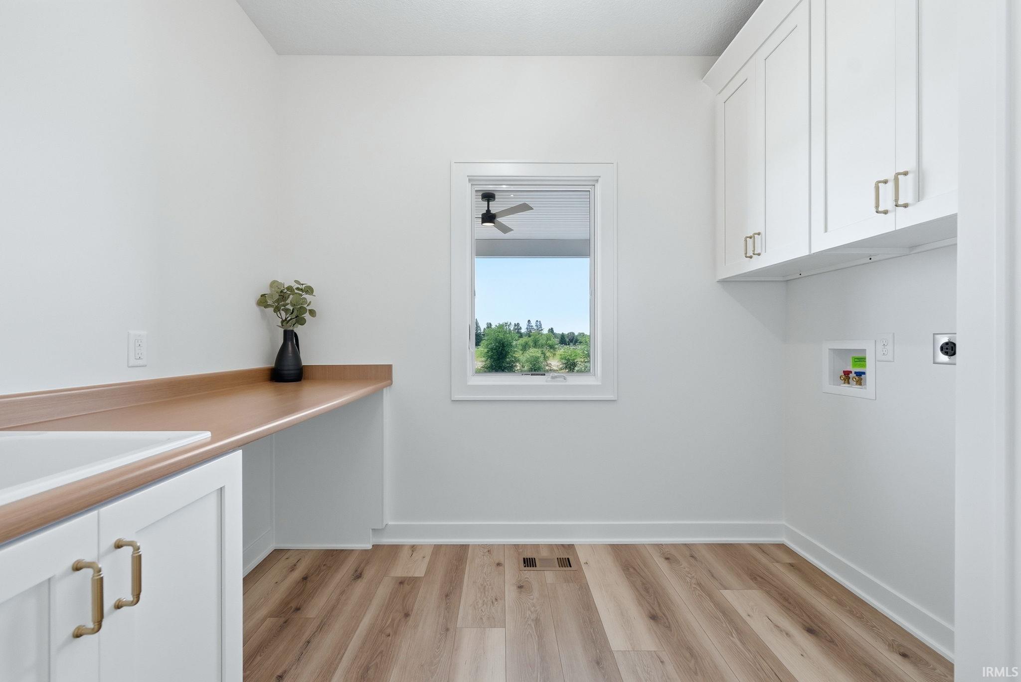 Laundry room featuring wood-finish flooring, white cabinetry with brass-toned hardware, a built-in sink with a wood-finish countertop, and a window providing natural light