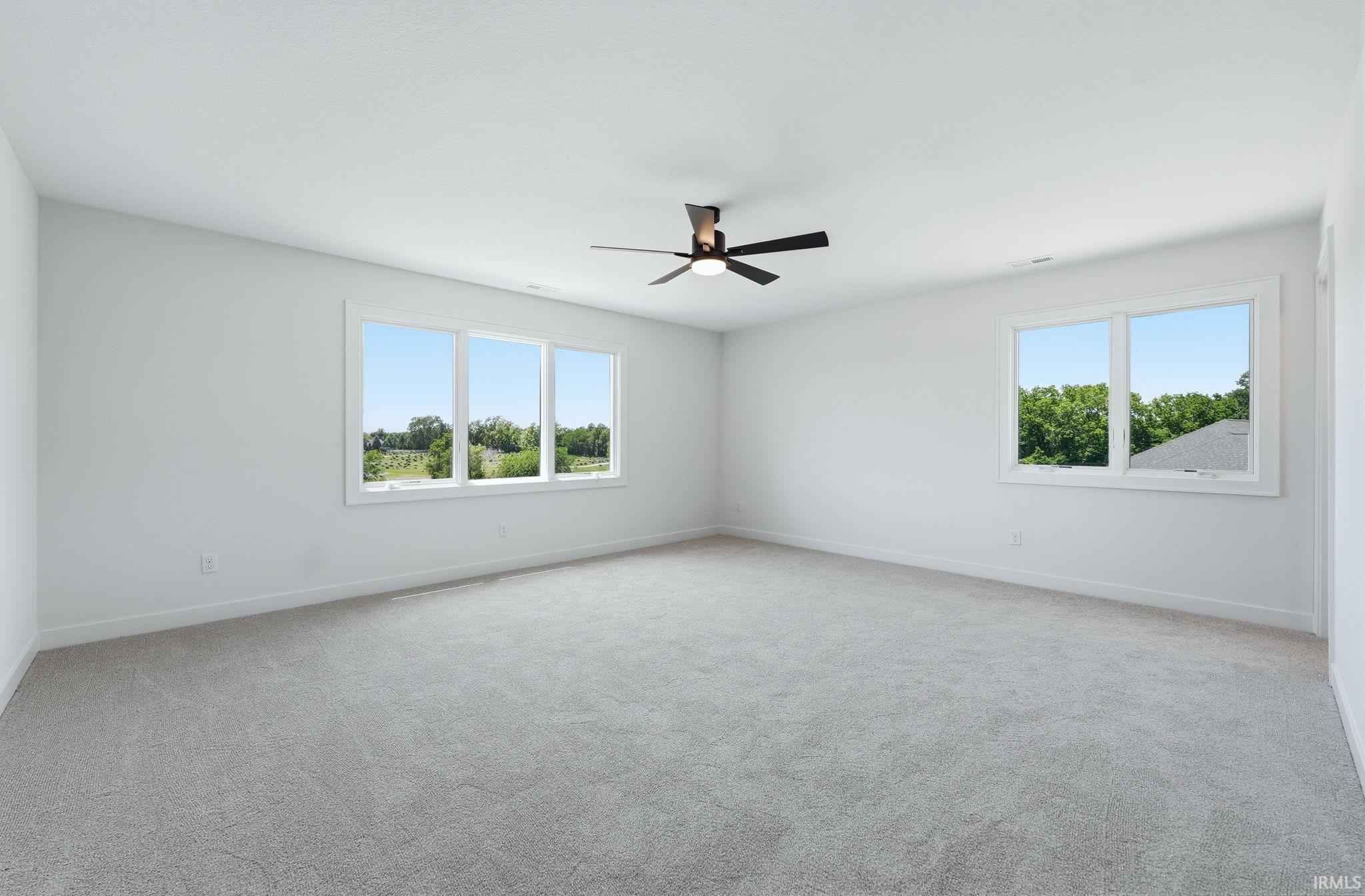 Spacious room featuring light gray carpeting, crisp white walls, multiple windows with white trim, a modern black ceiling fan with integrated lighting, and white baseboards