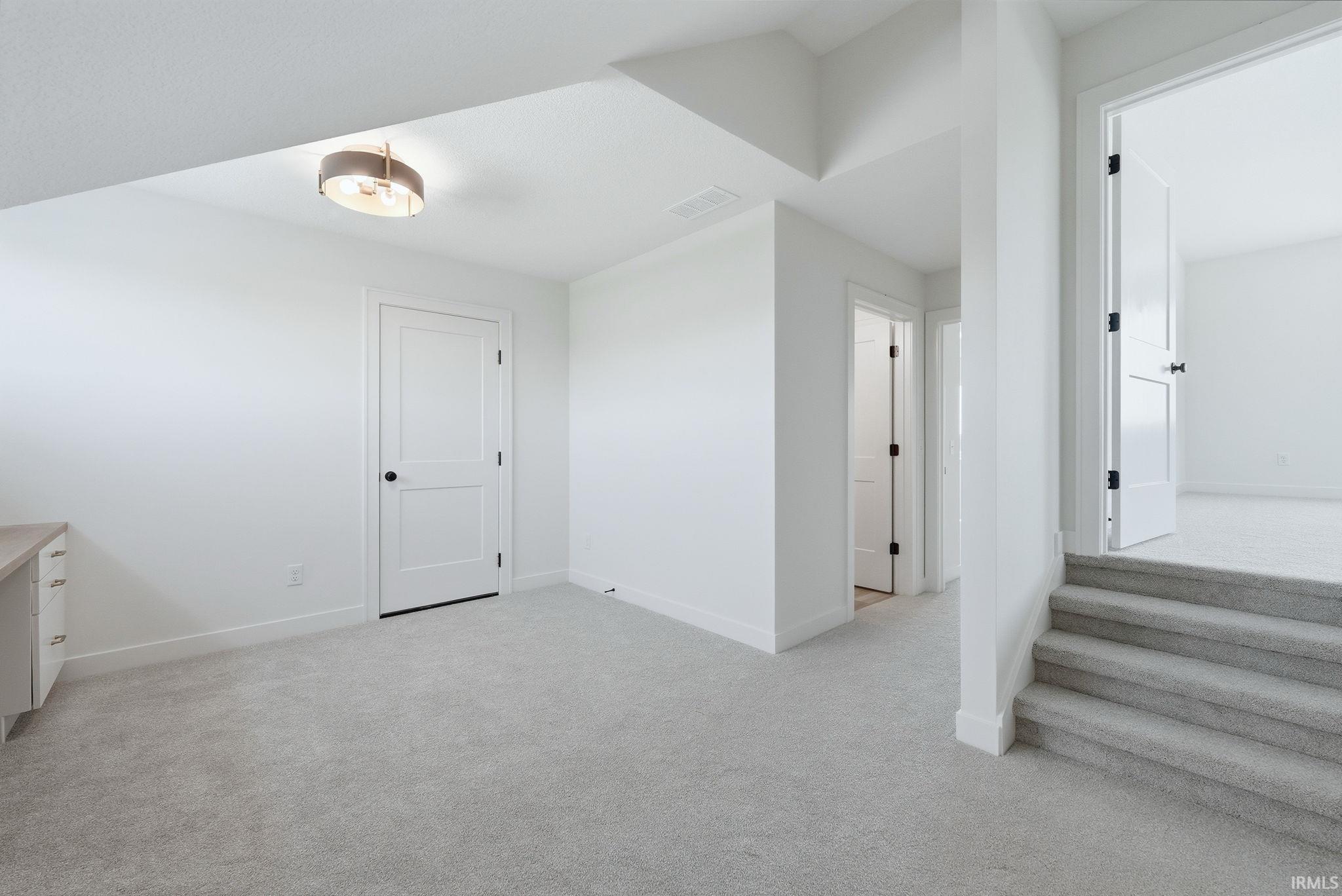 Carpeted interior space with white walls and trim, featuring a built-in desk with white cabinetry, a flush-mount ceiling light fixture, and multiple white panel doors with black hardware