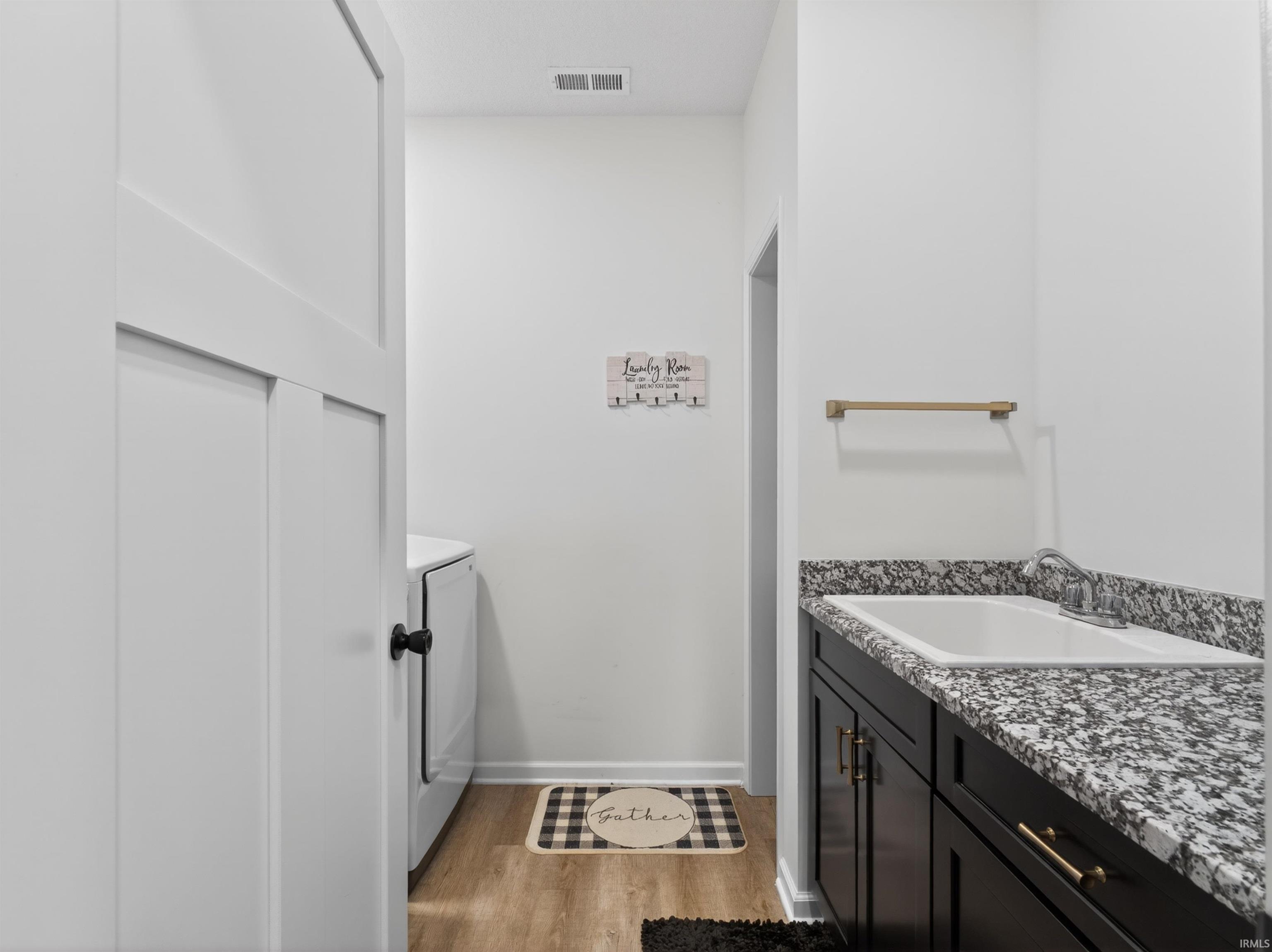 Laundry area featuring a utility sink with a granite-style countertop, dark cabinetry with brass hardware, light wood-finish flooring, and white walls