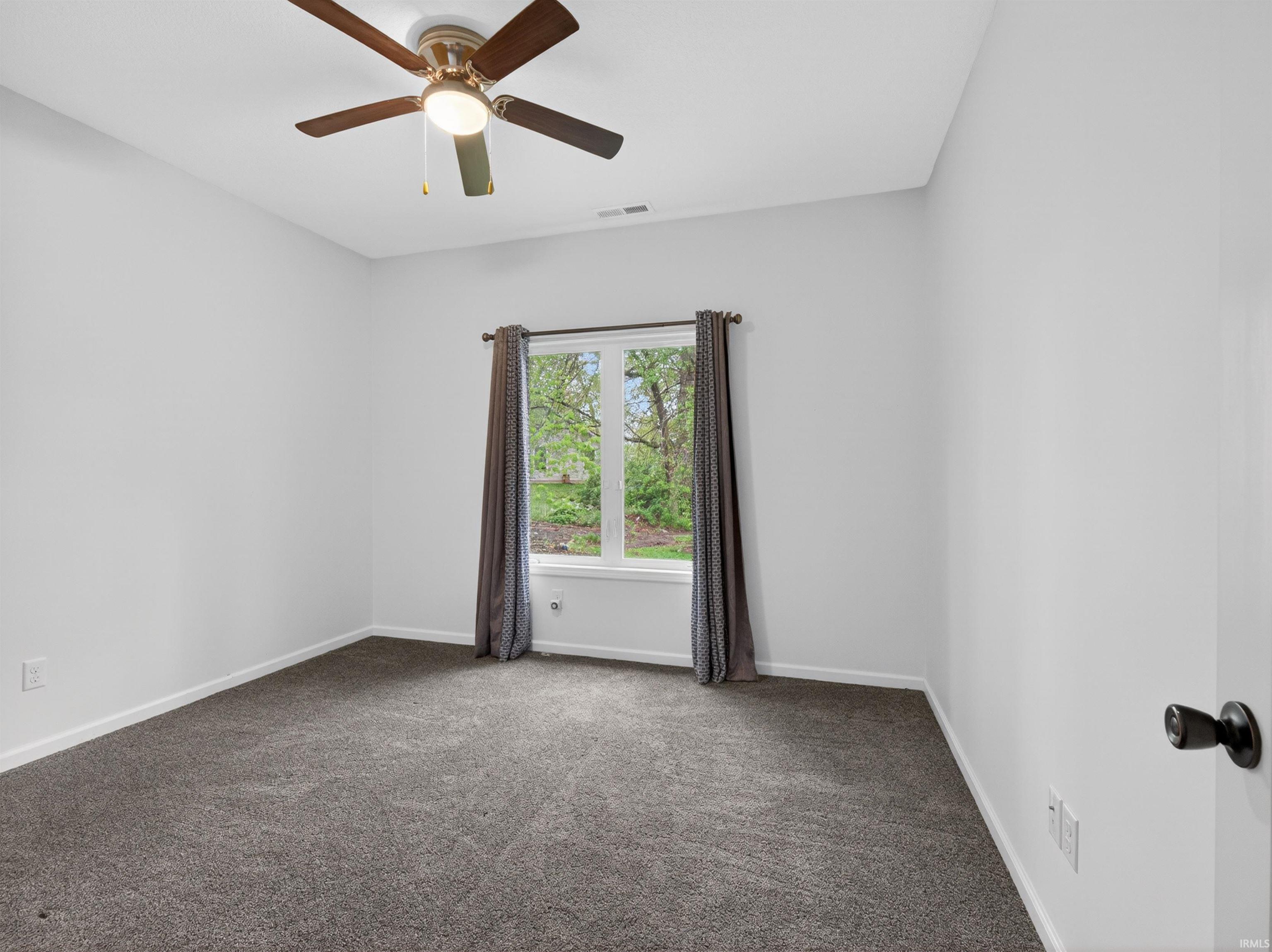 Neutral interior room featuring a window with exterior tree views, a ceiling fan with integrated lighting, and dark gray carpeting