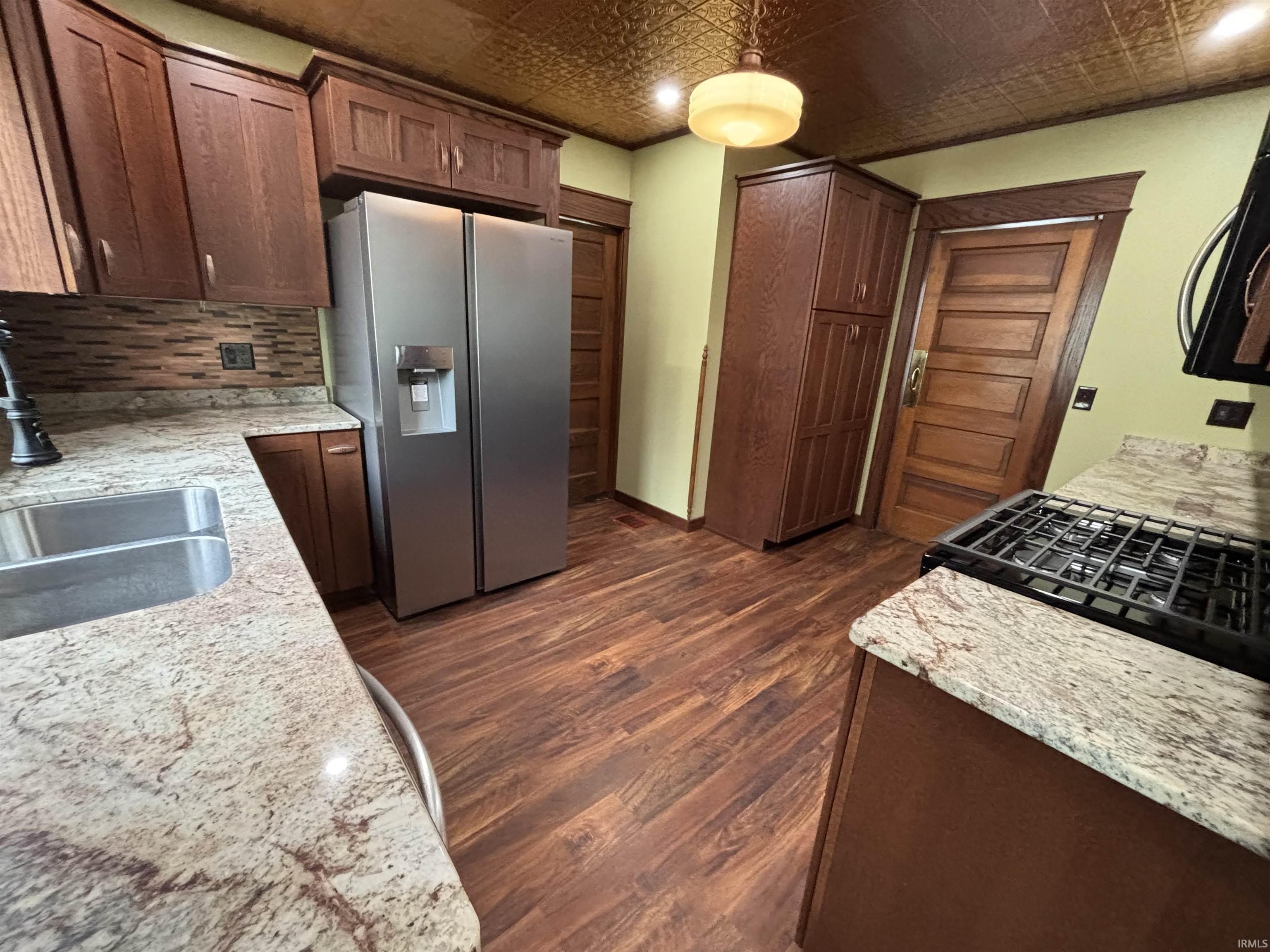 Kitchen featuring wood-finish flooring, granite-style countertops, stainless steel appliances, a tile backsplash, and dark wood cabinetry