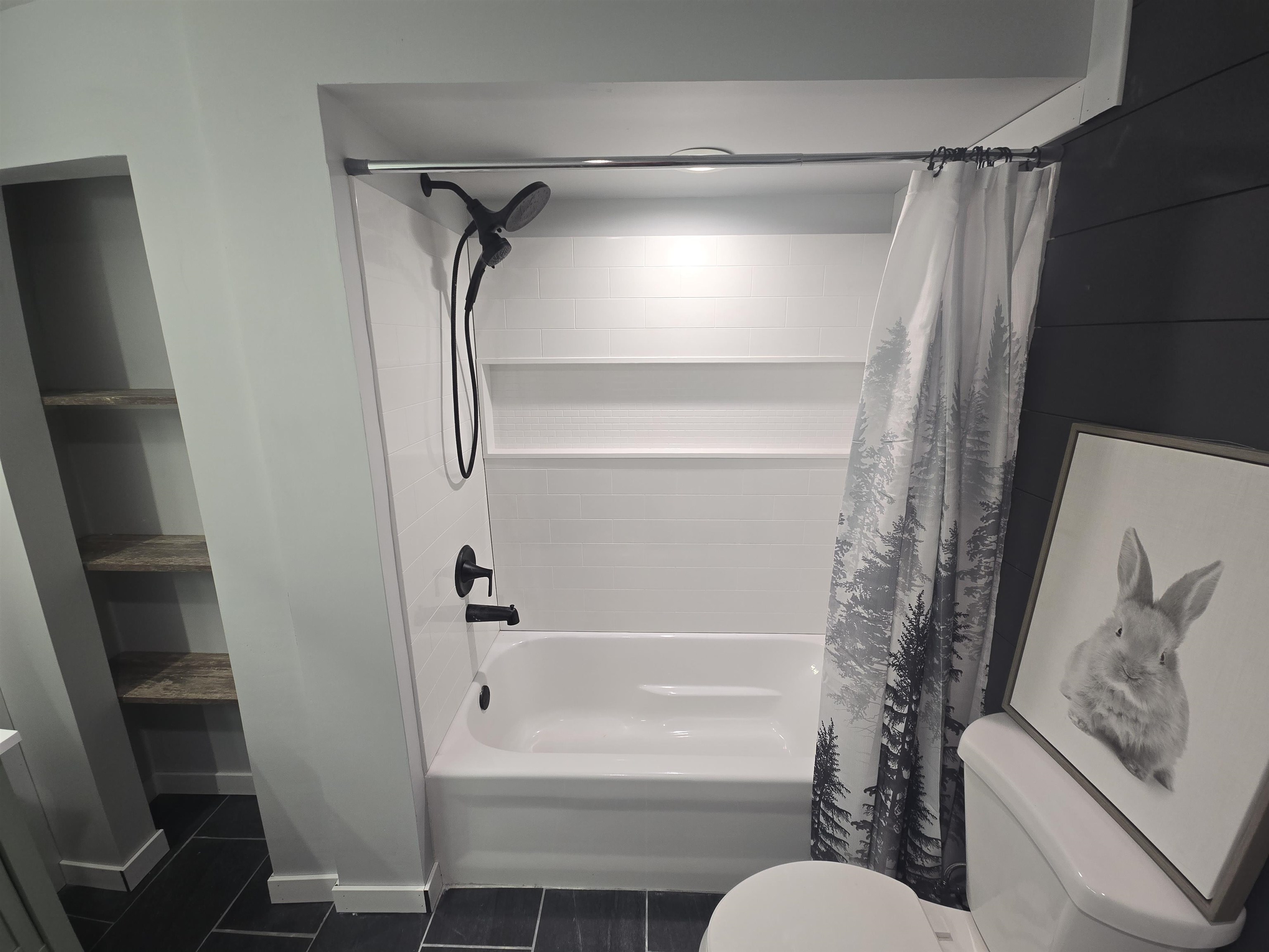 Bathroom featuring a white tub/shower combination with black fixtures, dark gray plank accent wall, and built-in shelving