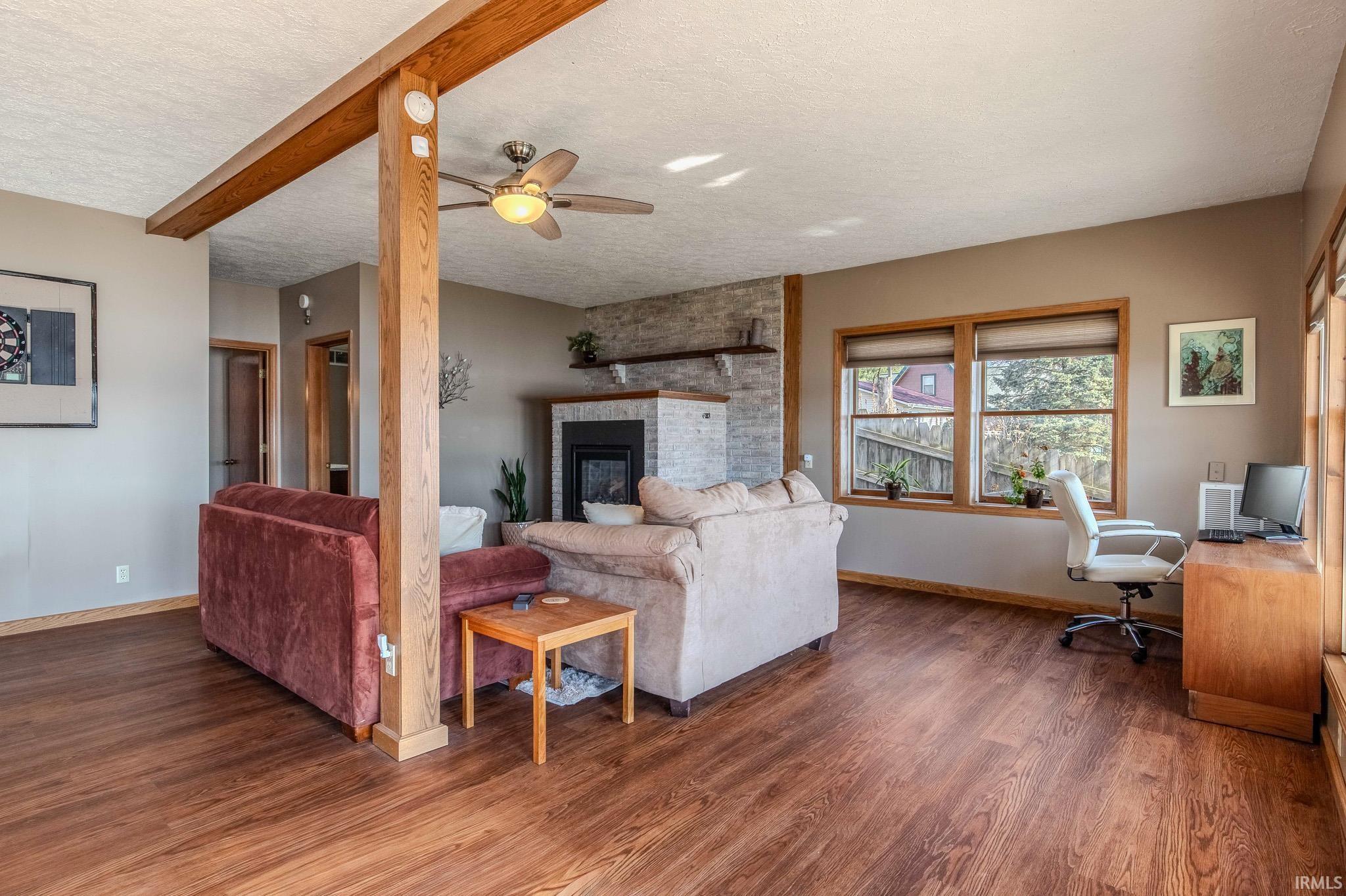 Spacious living area featuring wood-finish flooring, exposed wood beams, a stone-clad fireplace, and multiple windows with wood trim