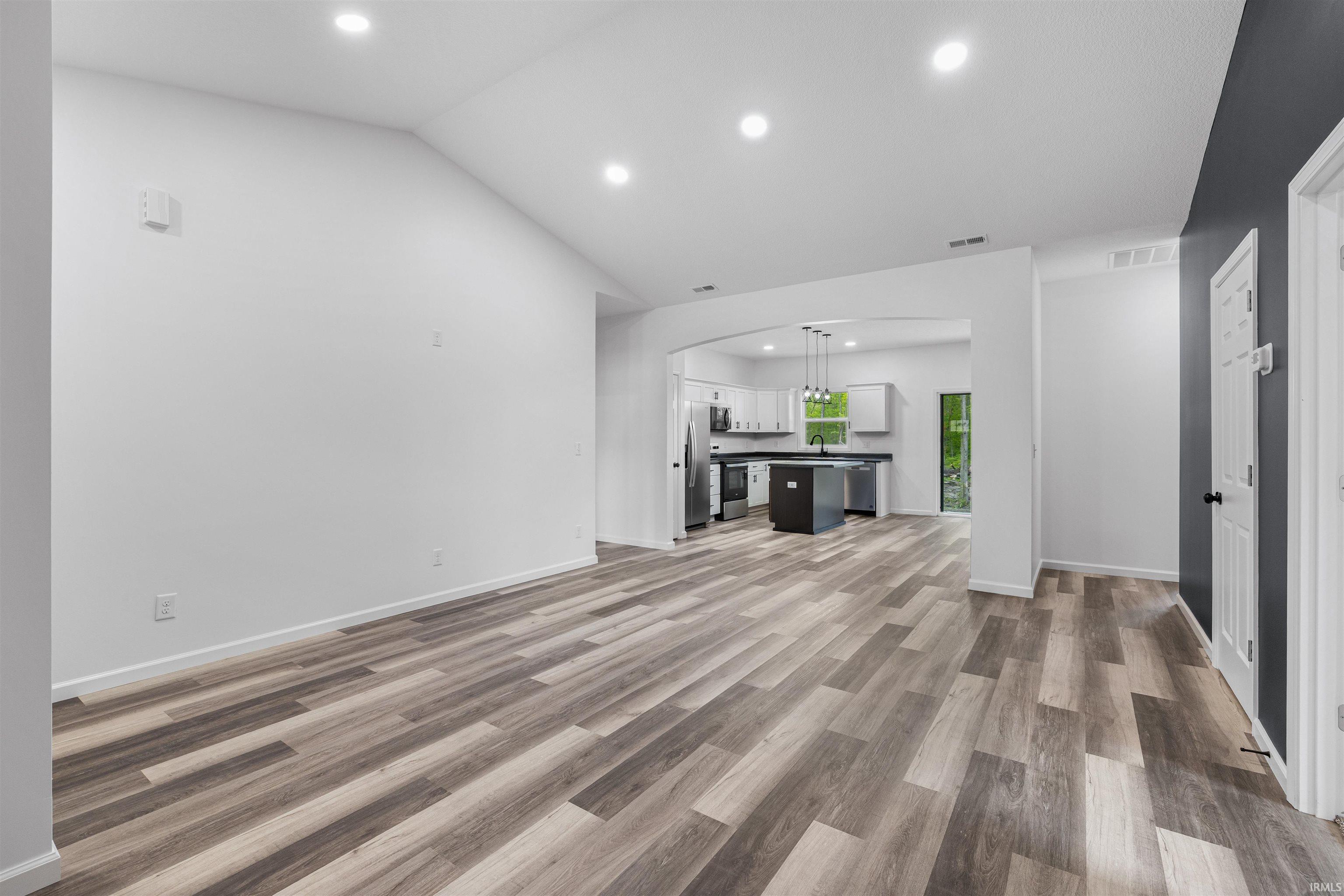 Spacious room featuring wood-finish flooring, vaulted ceilings with recessed lighting, and an open archway to the kitchen