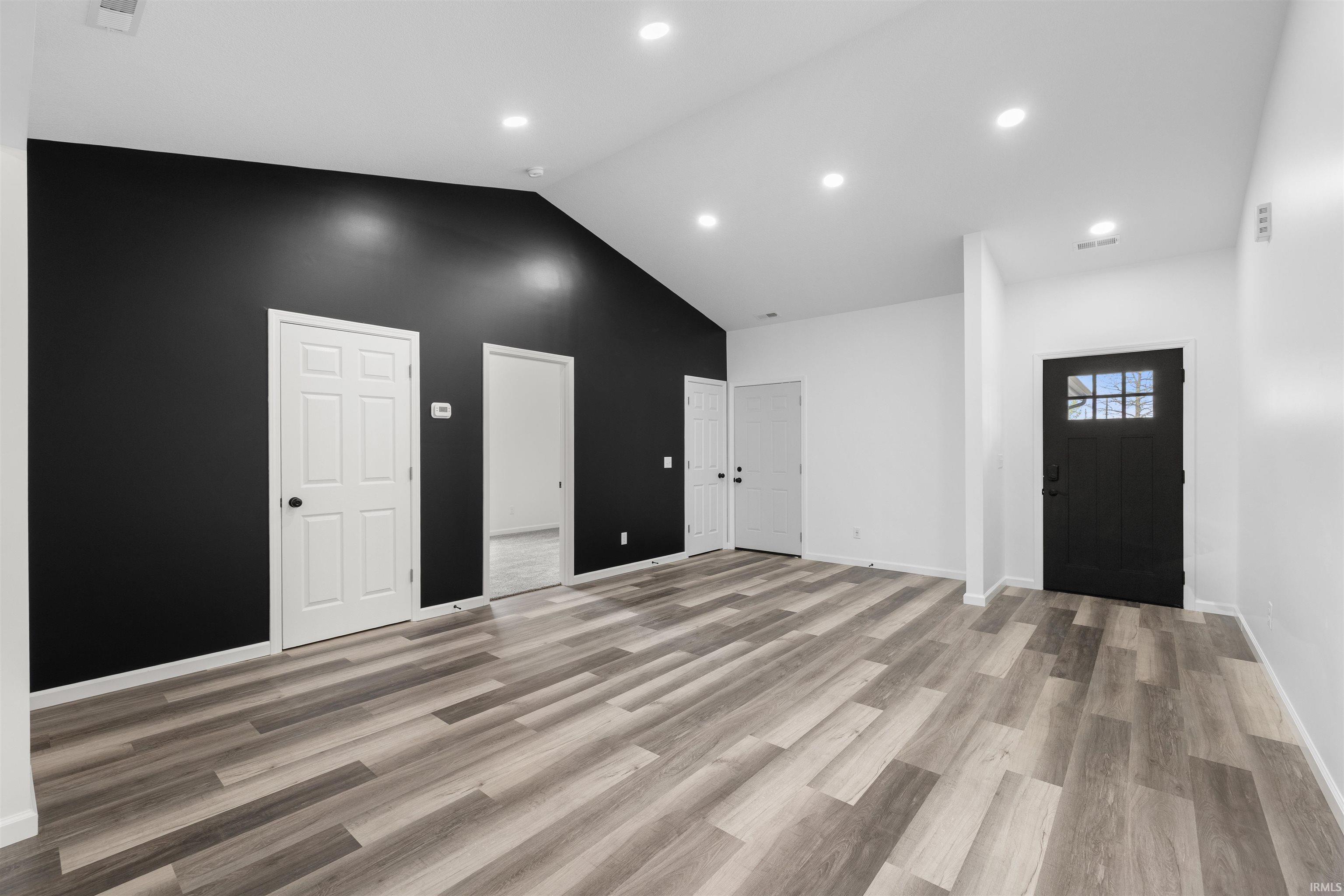 Spacious room featuring vaulted ceilings, recessed lighting, wood-finish flooring, a contemporary accent wall, and a craftsman-style entry door with divided lites