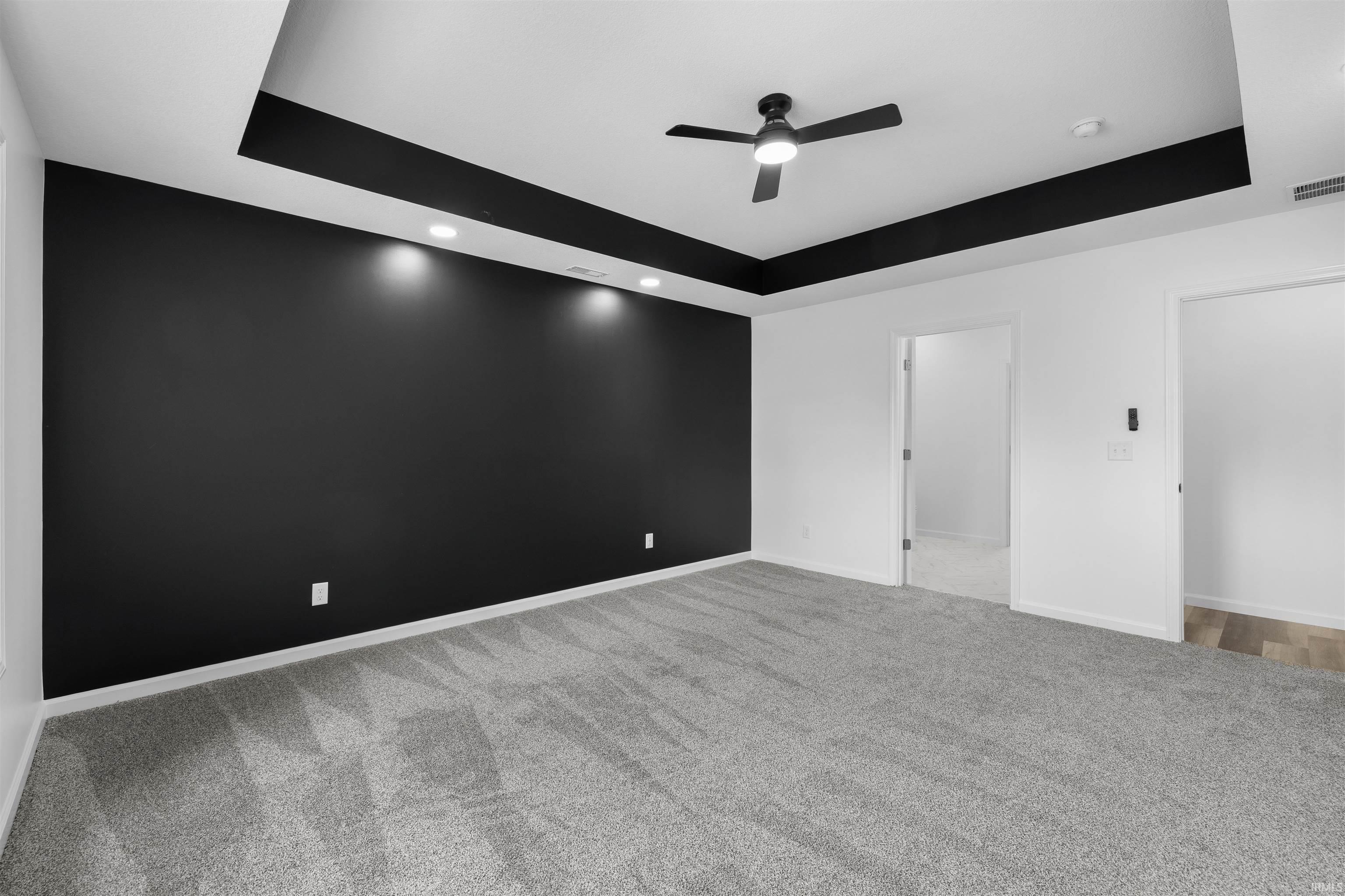 Spacious room featuring a dark accent wall with recessed lighting, a tray ceiling with dark trim, light gray patterned carpeting, and a ceiling fan