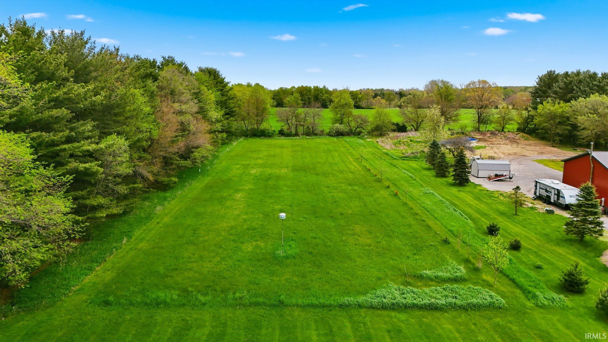 Expansive green lawn surrounded by mature trees, featuring a red barn structure, a gravel drive, and additional outbuildings