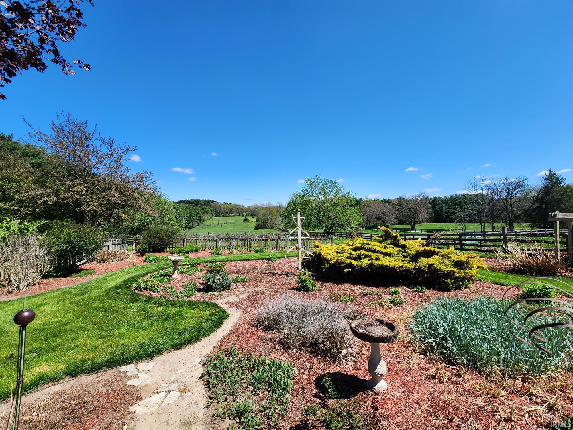 Expansive backyard garden featuring a manicured lawn path, varied plantings, decorative bird baths, and a wood fence bordering open fields