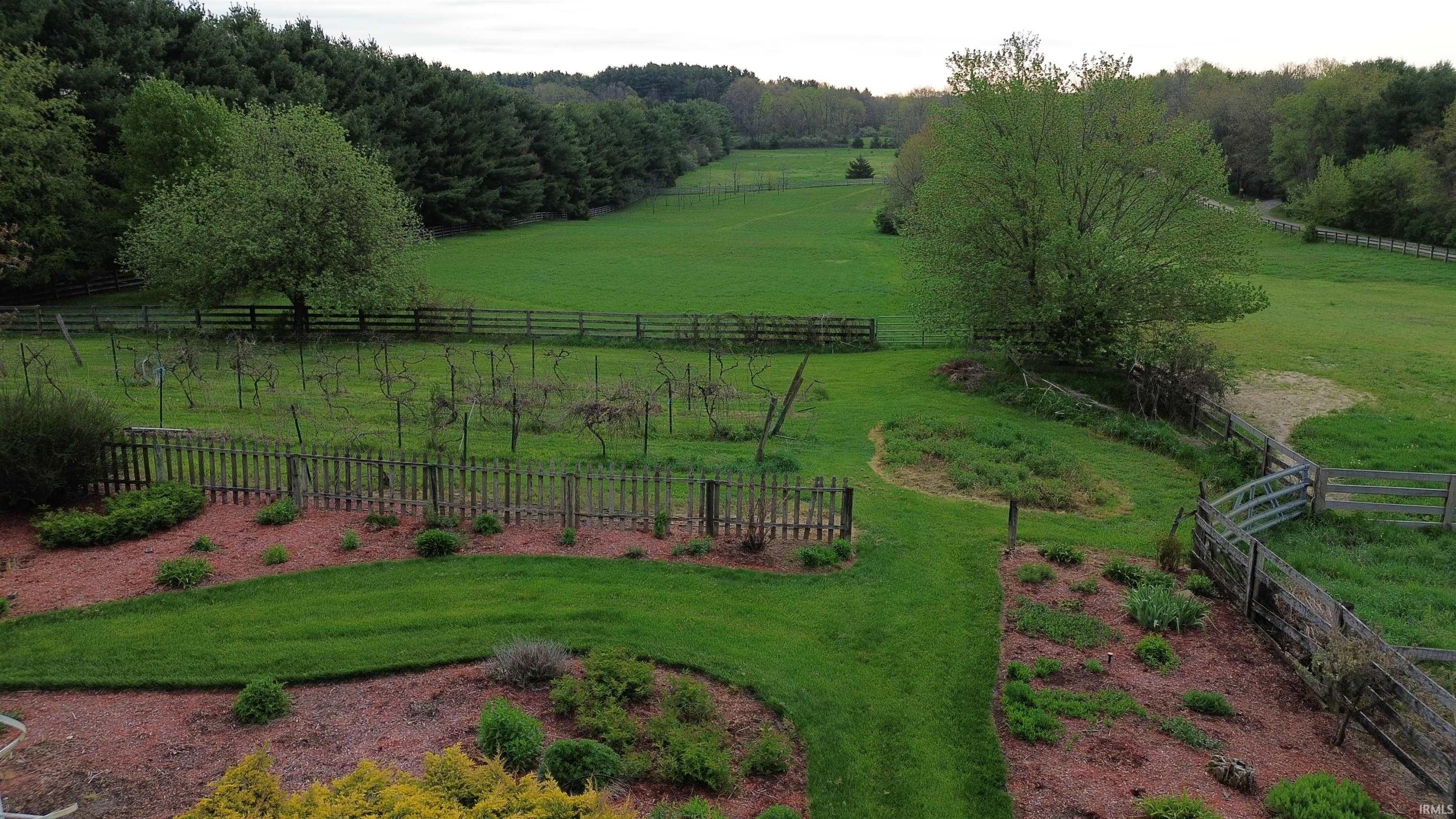 Expansive green space featuring fenced pastures, mature trees, landscaped beds with groundcover, and a defined grassy path