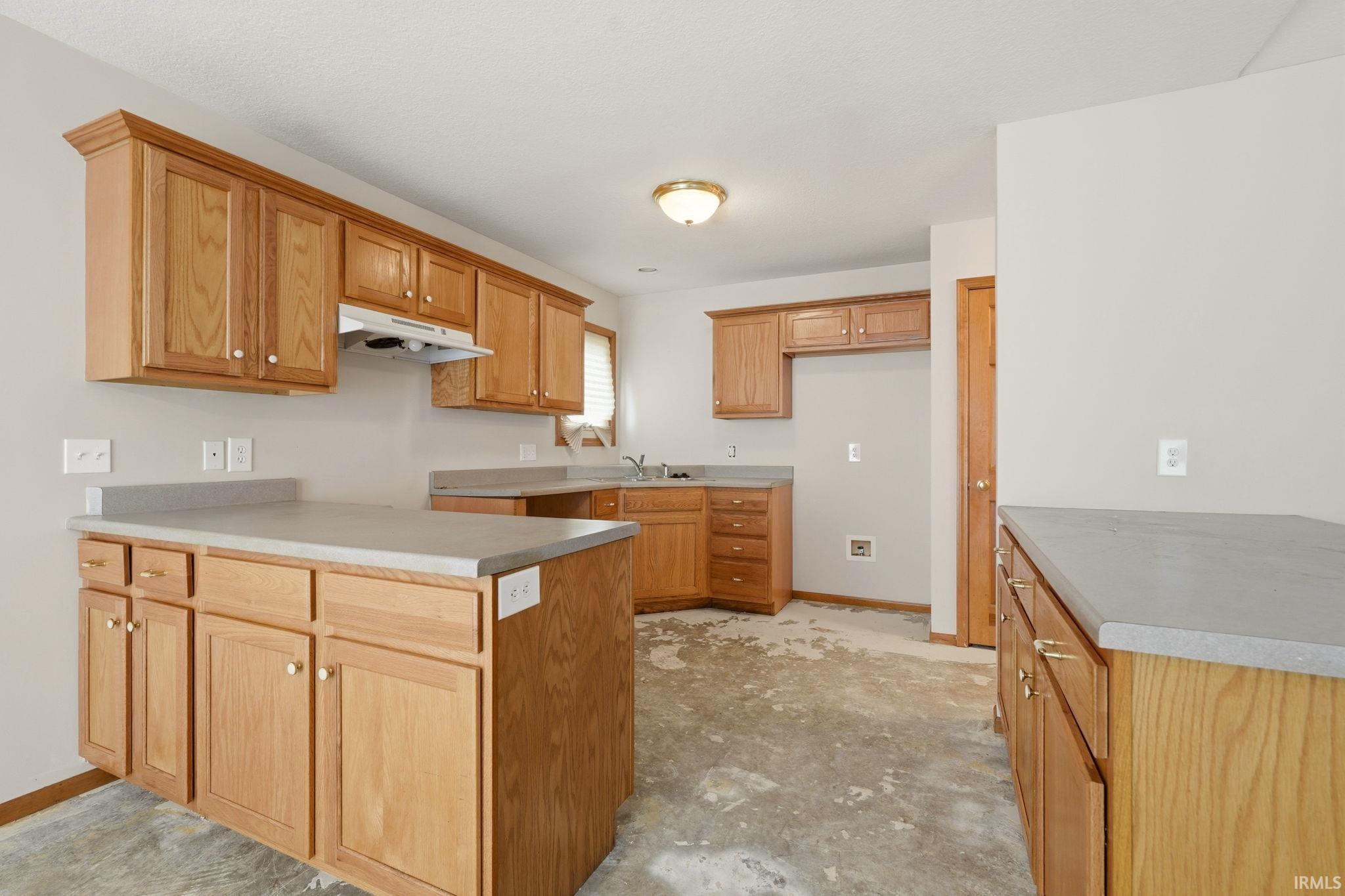 Kitchen featuring ample wood cabinetry, light-toned countertops, a built-in range hood, and a functional sink area with a window