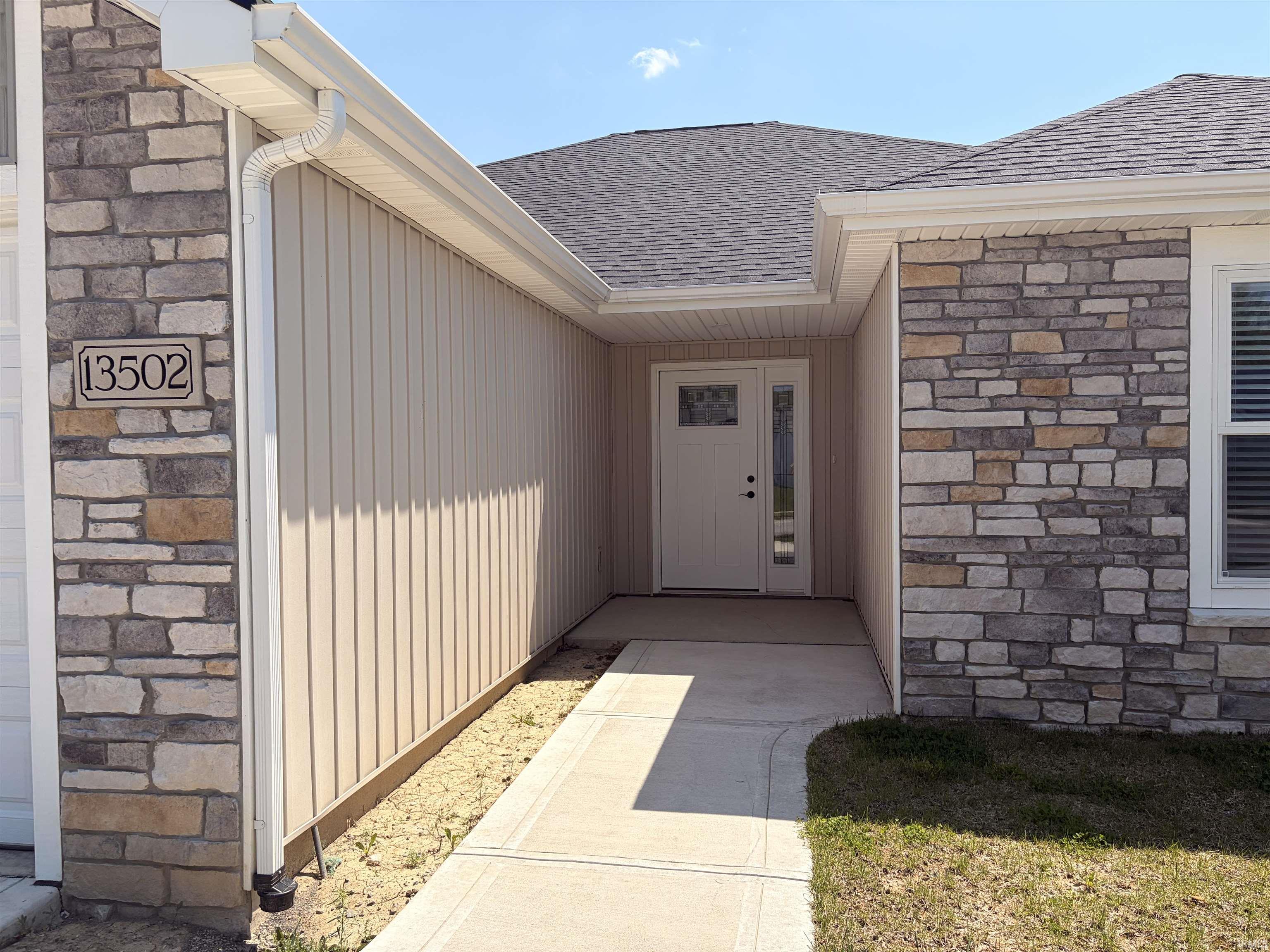Inviting front entry featuring a white paneled door with glass insert and sidelight, stone veneer facade, vertical siding, a paved walkway, and a shingled roof