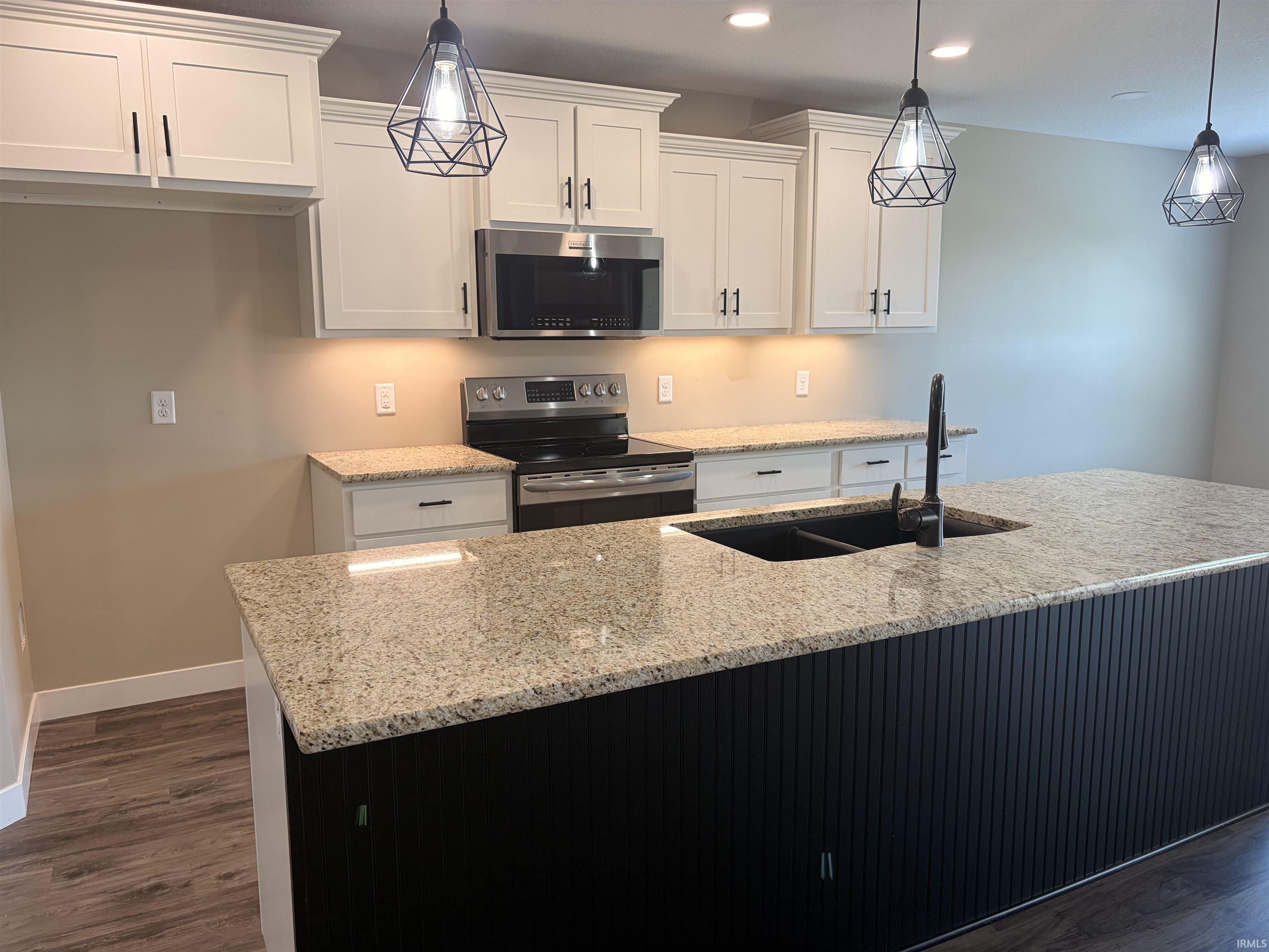 Kitchen featuring an expansive island with a dual basin sink and dark textured paneling, light-toned countertops, white shaker-style cabinetry, stainless steel appliances, and wood-finish flooring
