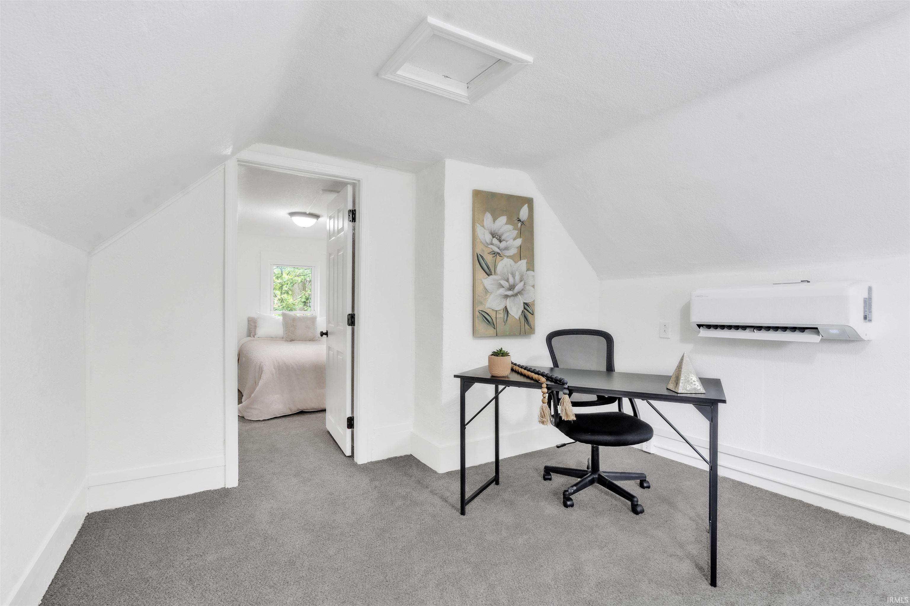 Carpeted room with sloped ceilings featuring a wall-mounted air conditioning unit, white baseboards, and a doorway opening to another room with a window