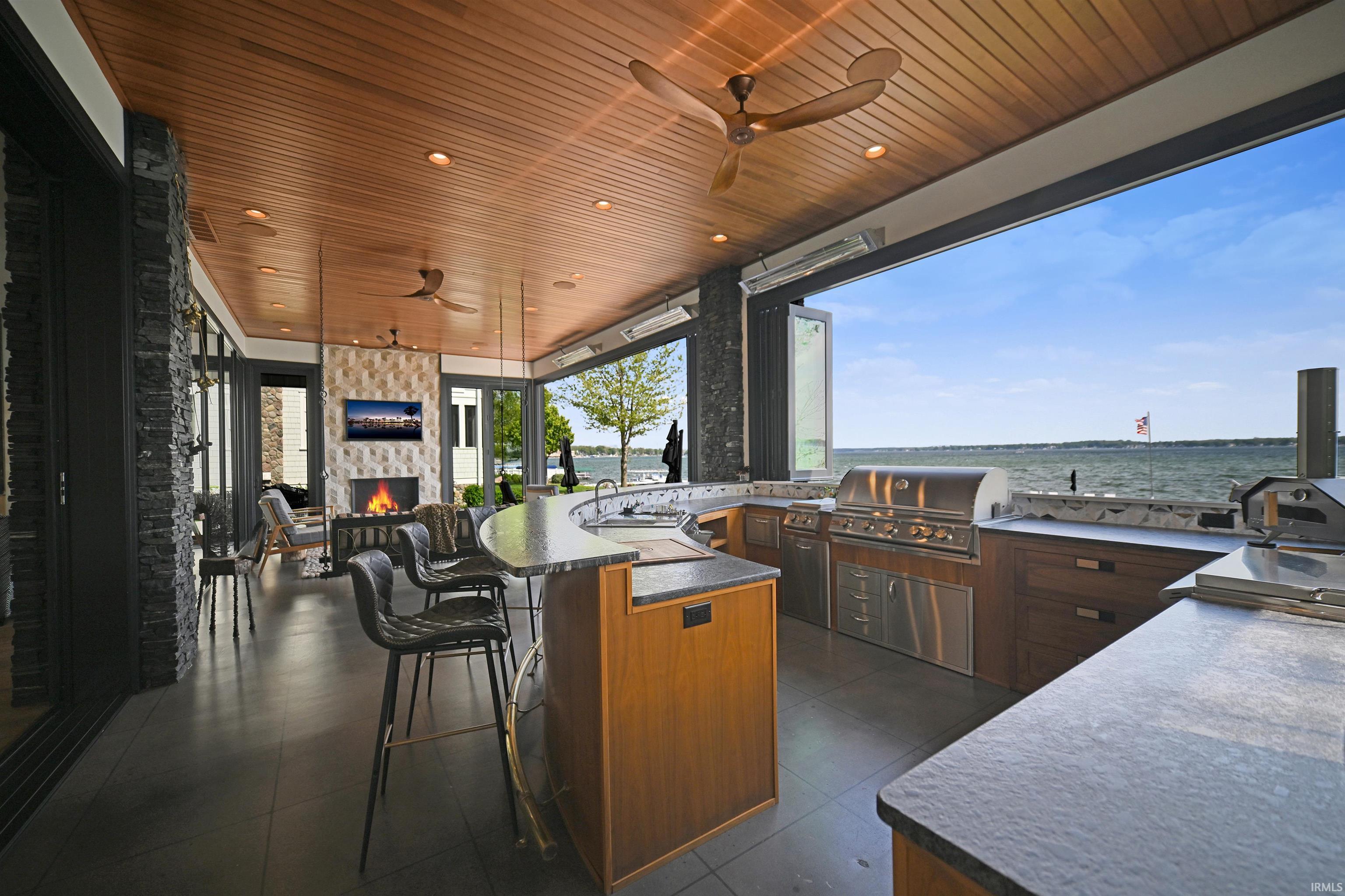 Open-air living space featuring a wood-finish ceiling, built-in outdoor kitchen with stainless steel appliances, curved bar seating, and a stone-clad fireplace