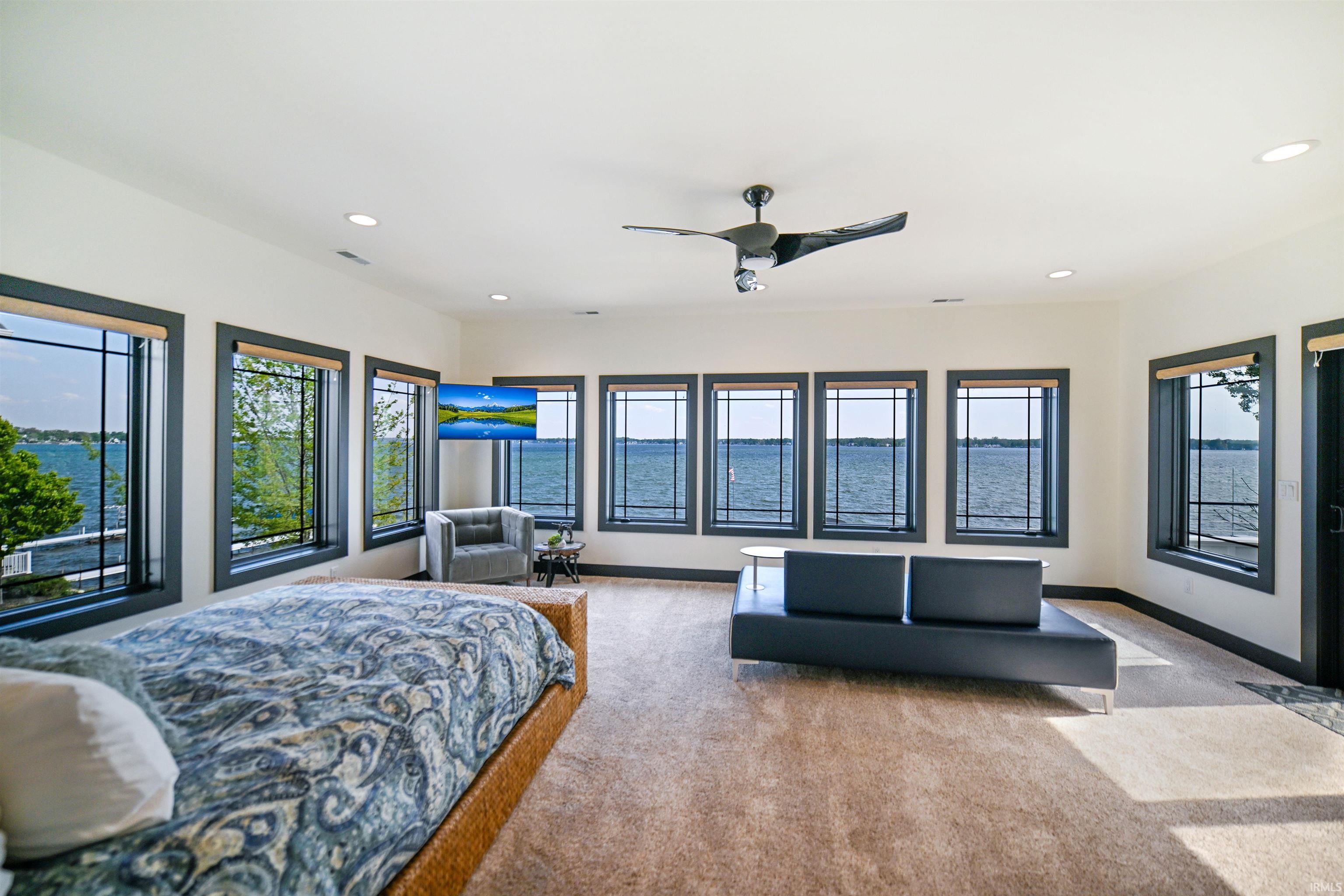 Spacious room featuring broadloom carpeting, a ceiling fan, recessed lighting, and numerous large windows with dark trim overlooking a body of water