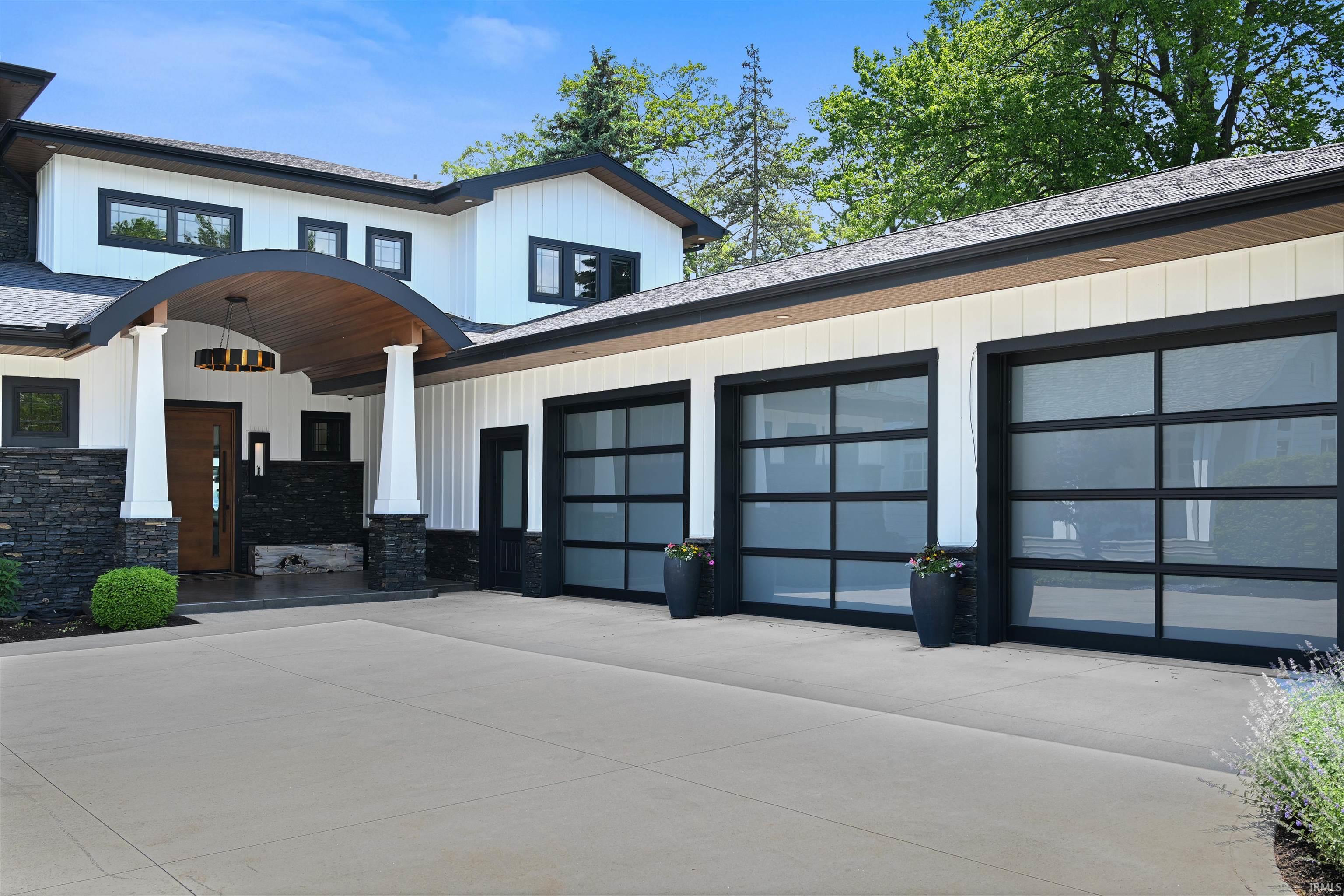 Contemporary white exterior with black trim, three frosted glass garage doors, a curved wood-finish entry canopy, black stone accents, and a light-colored driveway