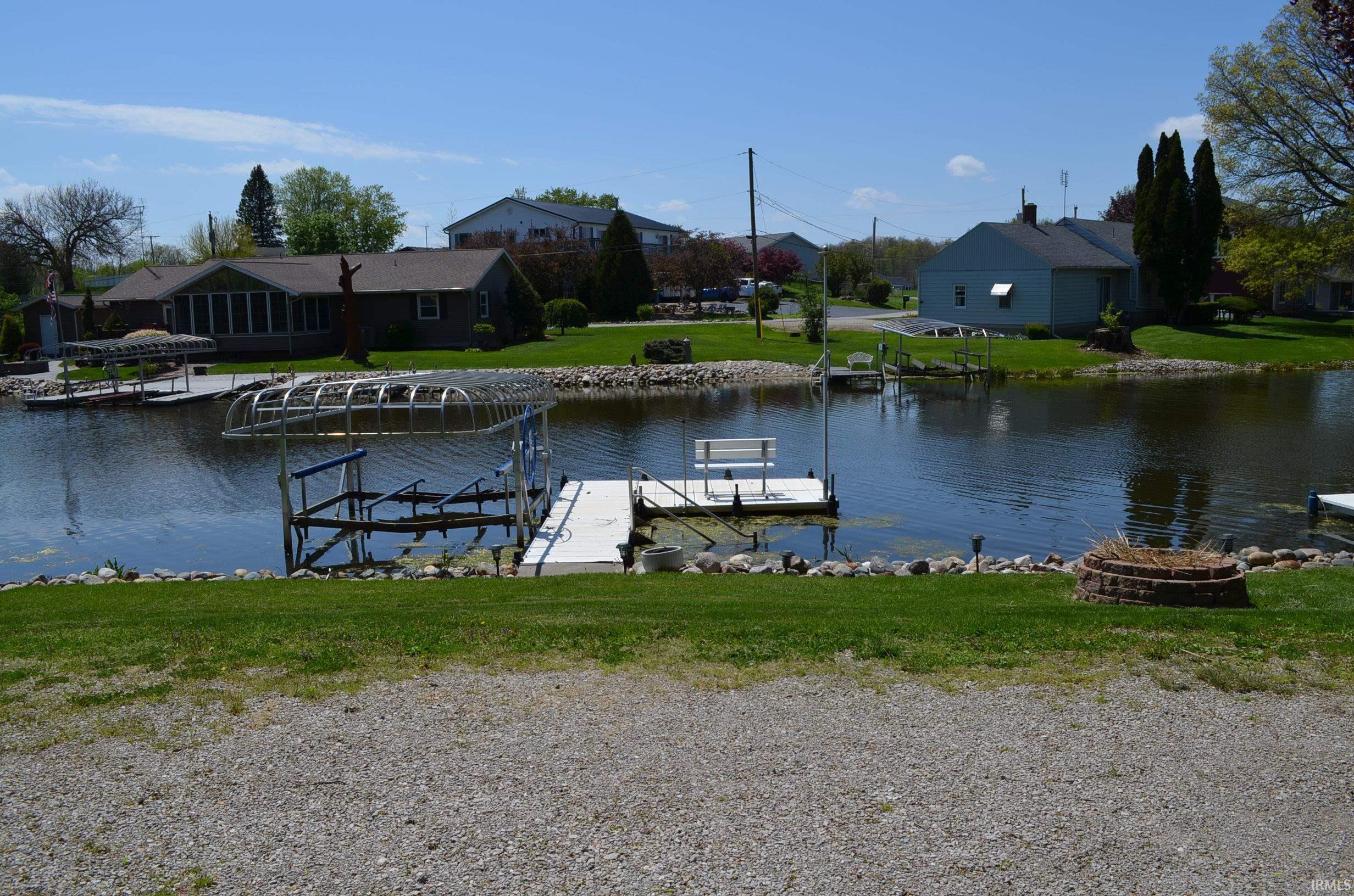 Waterfront property featuring a private boat lift, a dock with a bench, a gravel driveway, a stone-lined fire pit, and landscaped grassy areas