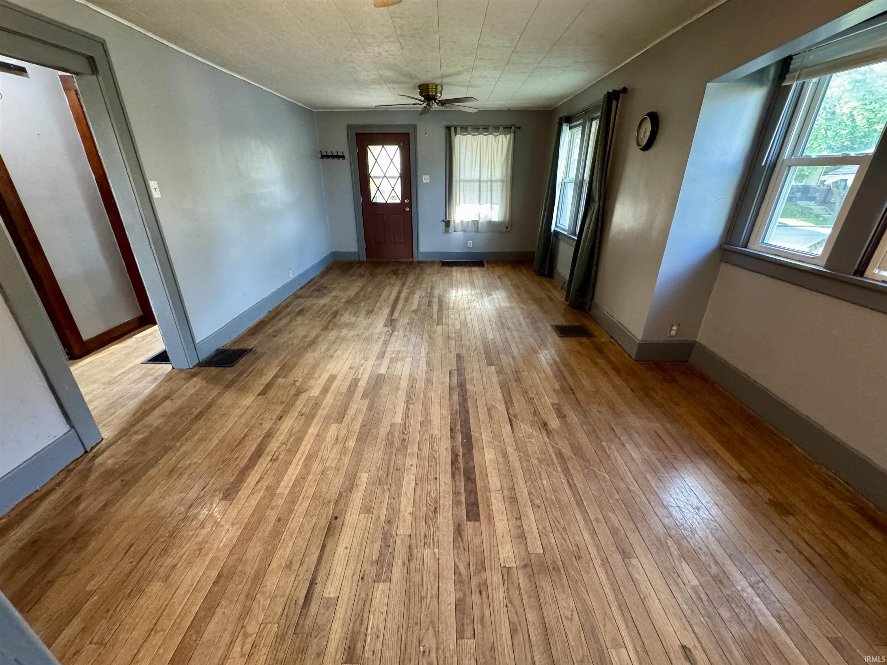 Spacious room featuring hardwood flooring, multiple windows, a ceiling fan, and a paneled entry door