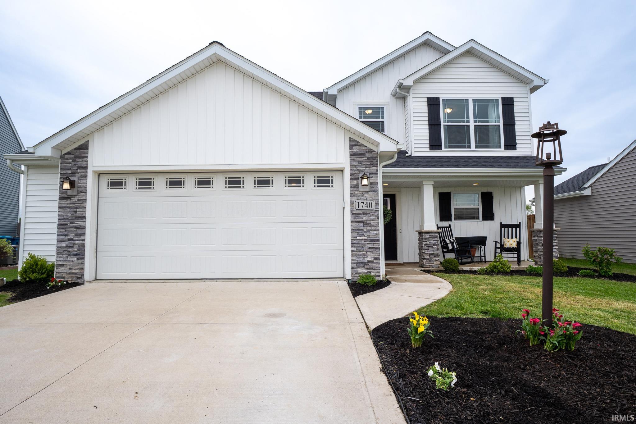 Two-story residence featuring white siding and dark shutters, a two-car garage with transom windows, a covered front porch with stone-accented pillars, a concrete driveway, and landscaped beds with mulch and plantings