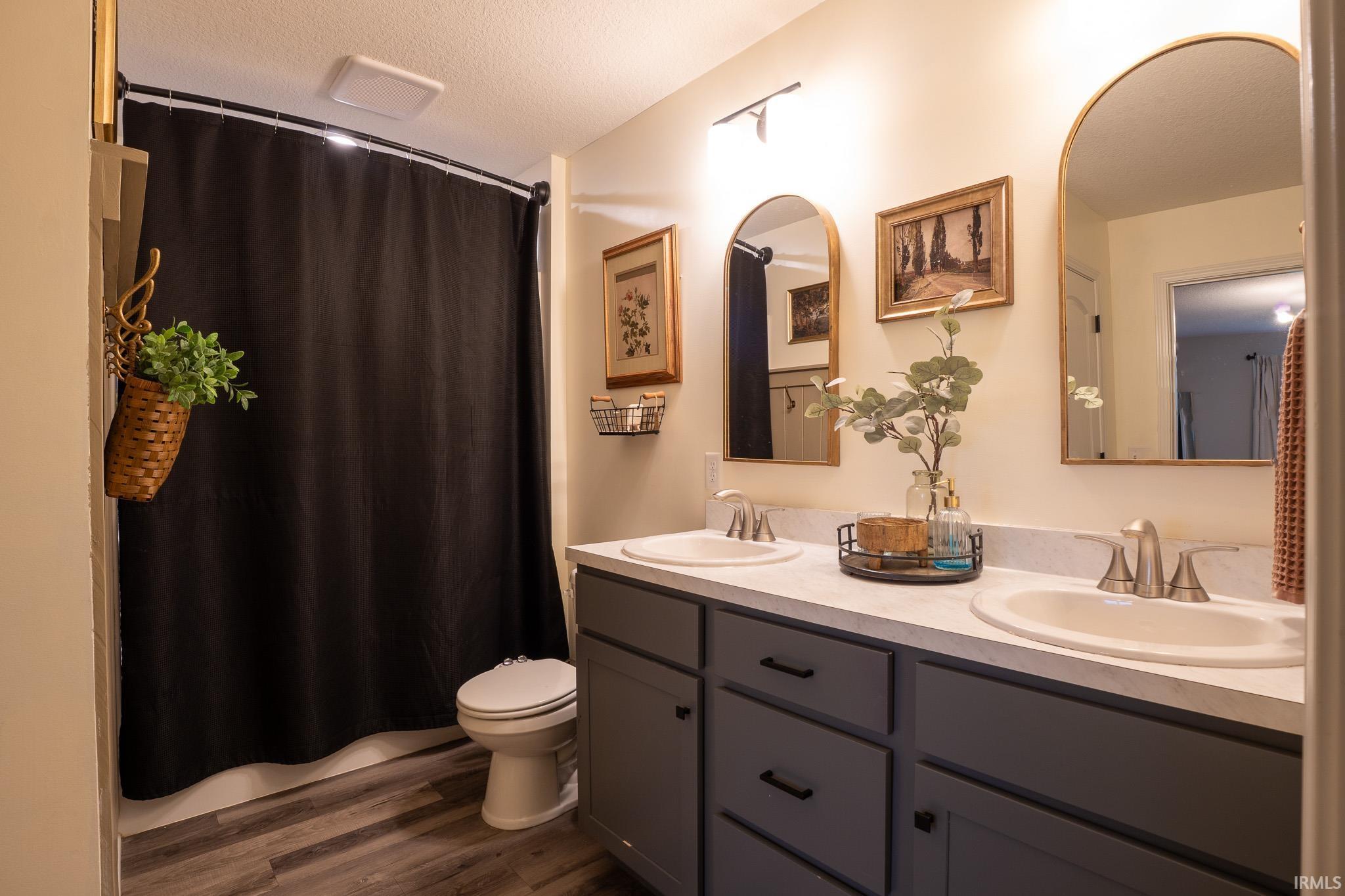 Bathroom featuring a dual vanity with undermount sinks, wood-finish flooring, a toilet, and a shower area with a dark curtain