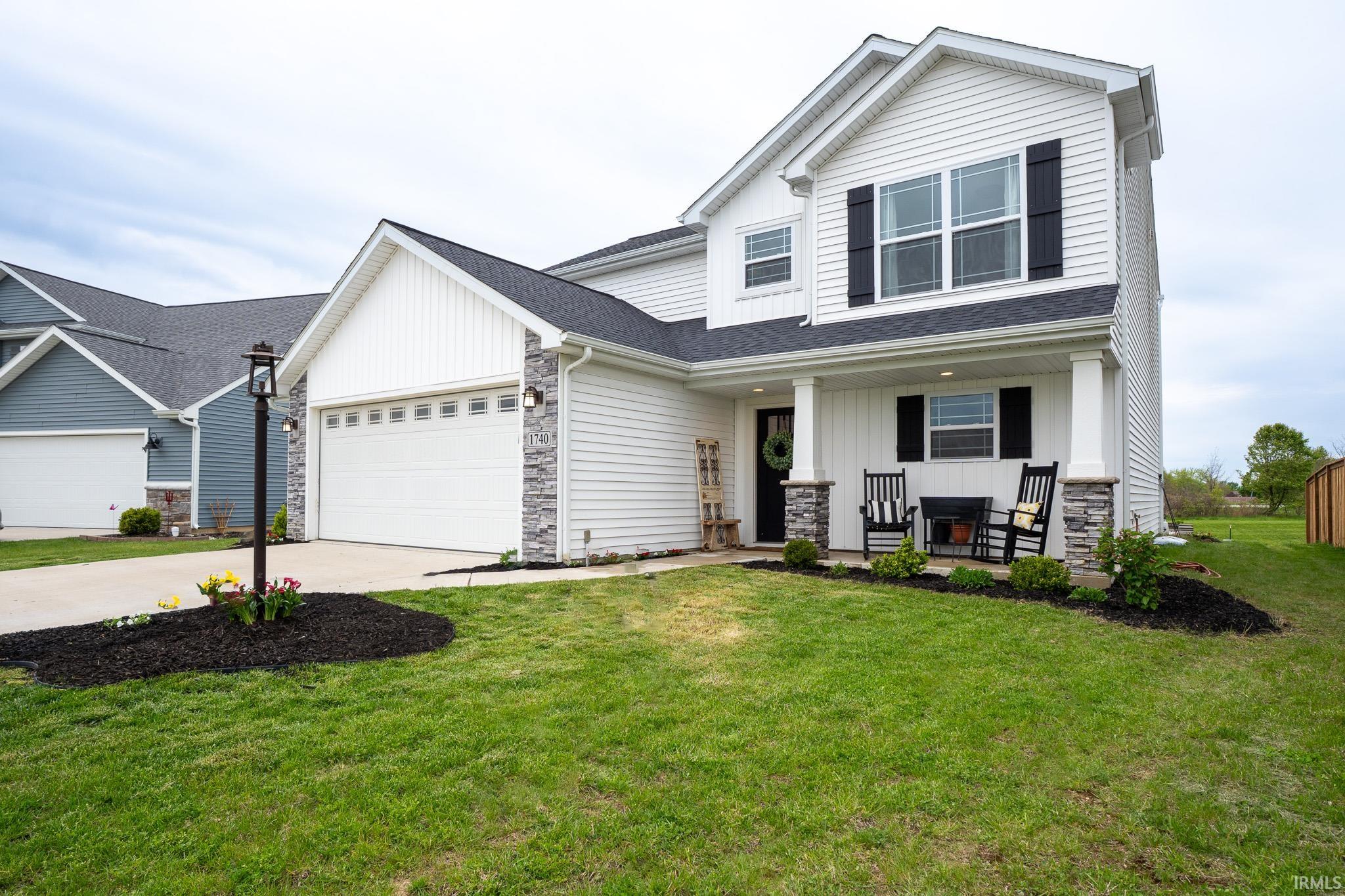 Two-story white siding exterior featuring a covered front porch with stone-accented pillars, black window shutters, a two-car garage, and a concrete driveway
