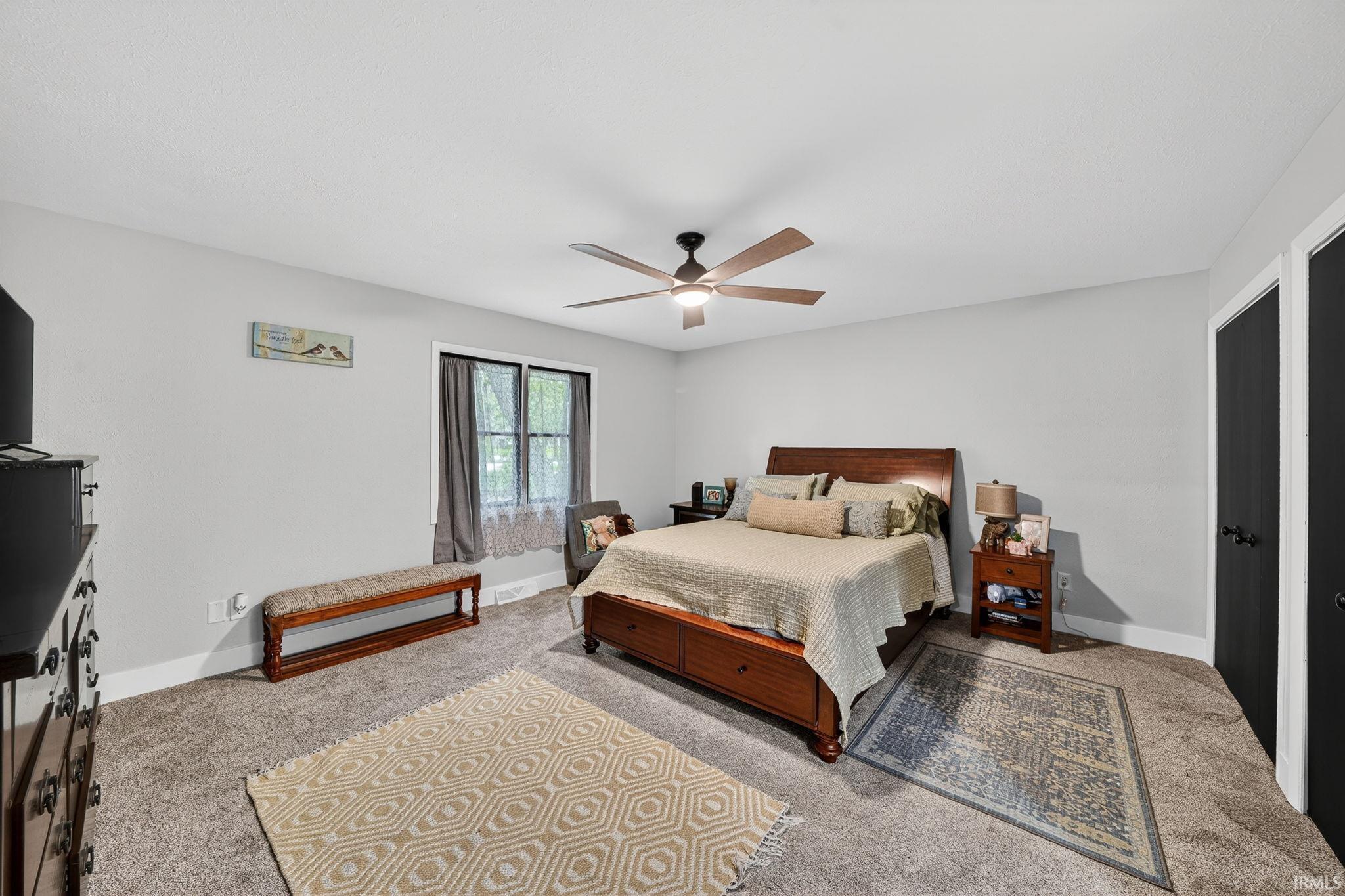 Spacious bedroom featuring light gray walls, carpet flooring, a multi-pane window with natural light, a ceiling fan with light fixture, and two black doors with dark hardware