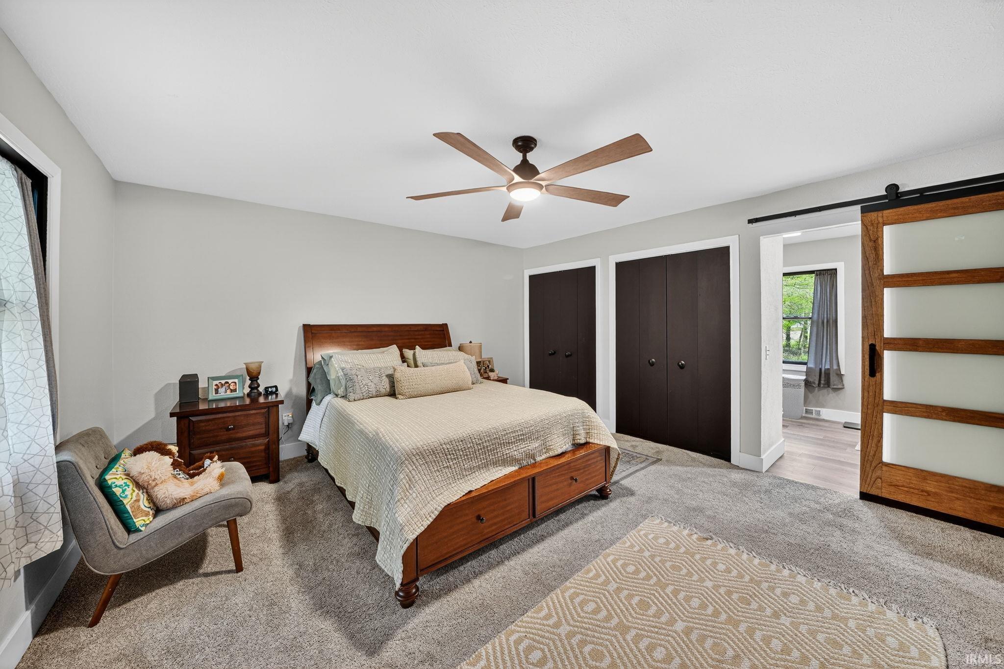 Spacious bedroom featuring light gray wall paint, gray carpeting, a multi-blade ceiling fan with integrated lighting, and two dark-paneled bifold closet doors