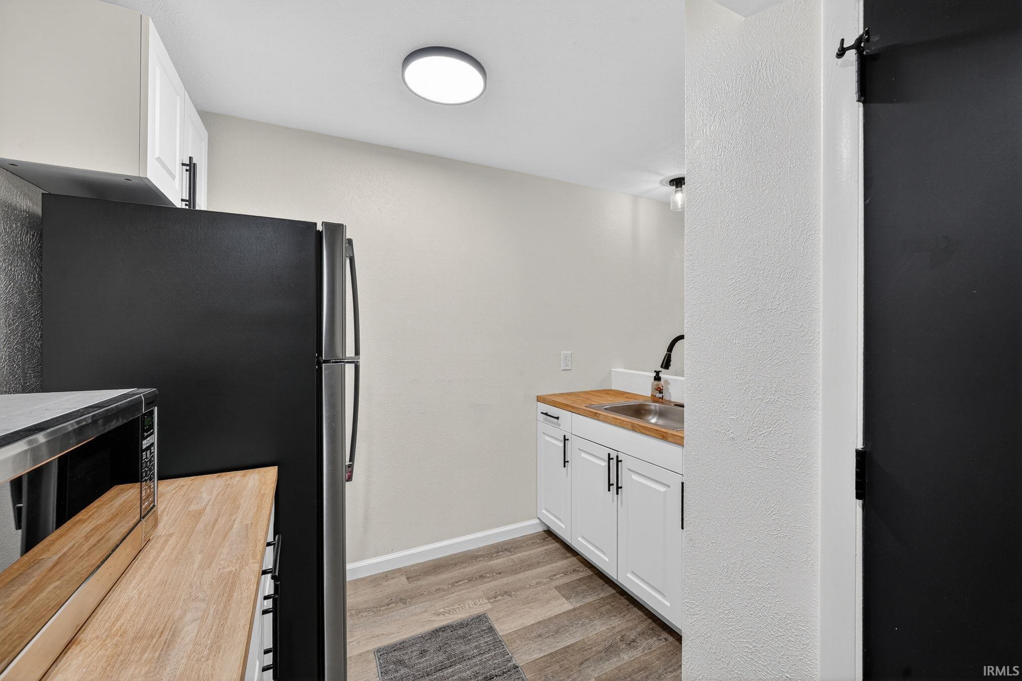 Compact kitchen area featuring wood-finish flooring, white cabinetry with dark hardware, a stainless steel sink with a dark faucet, a black refrigerator, and an overhead flush-mount light fixture