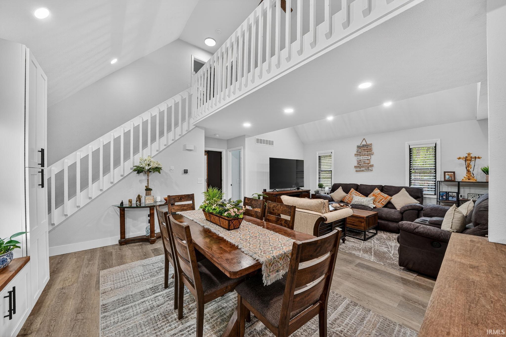 Open-concept living space featuring wood-finish flooring, recessed lighting, a vaulted ceiling, and a prominent staircase with white balusters