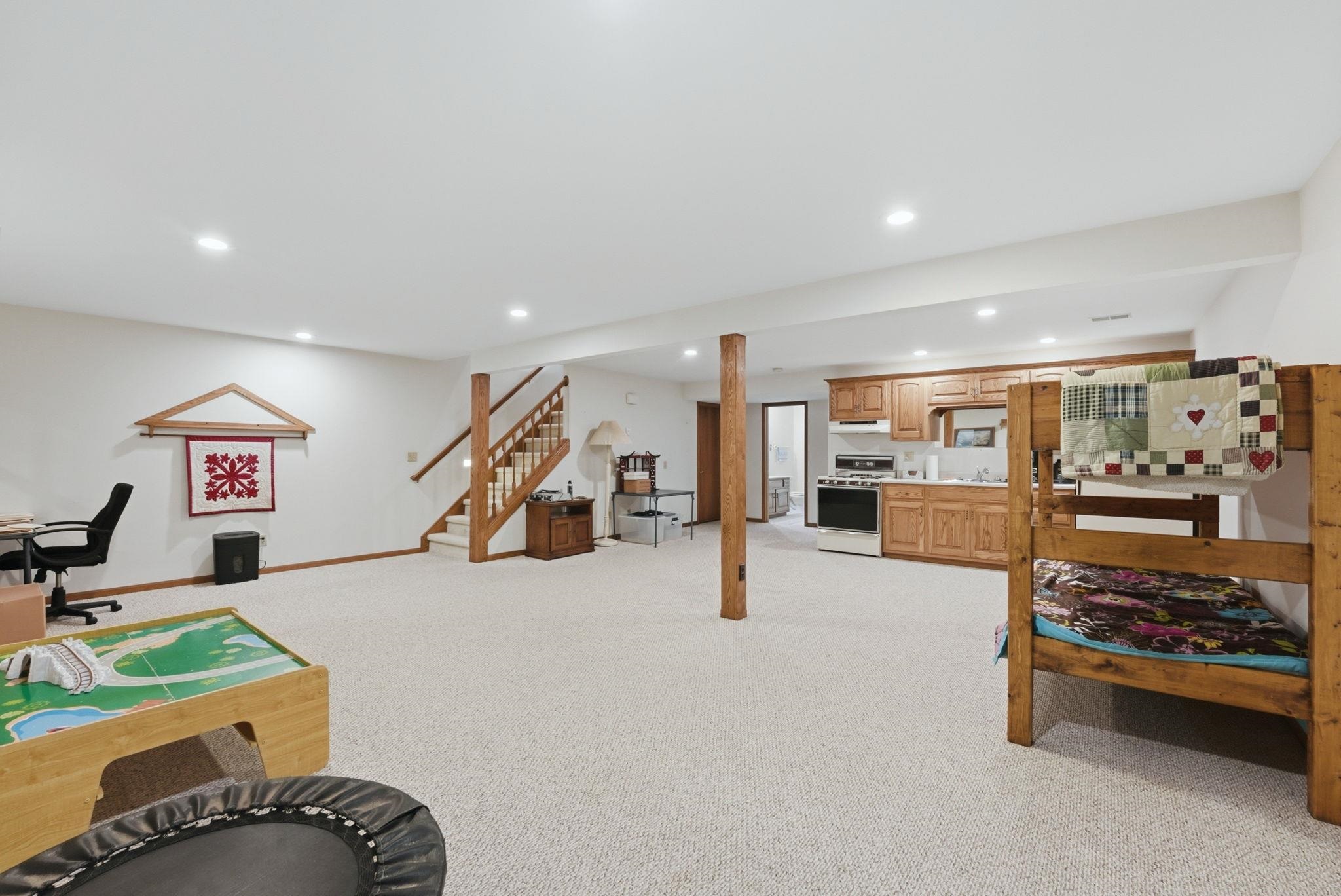 Expansive carpeted lower level featuring recessed lighting, a built-in kitchenette with wood cabinetry and white appliances, and a wood staircase with white risers