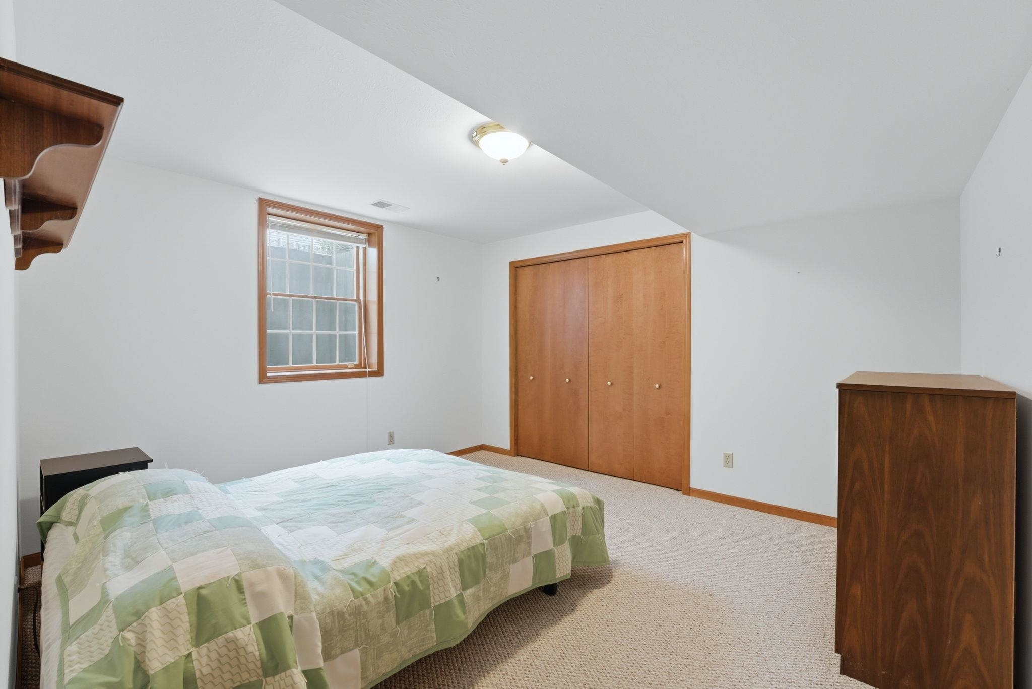 Carpeted room with a double-door closet featuring wood-finish doors, a window with wood trim, and a ceiling-mounted light fixture