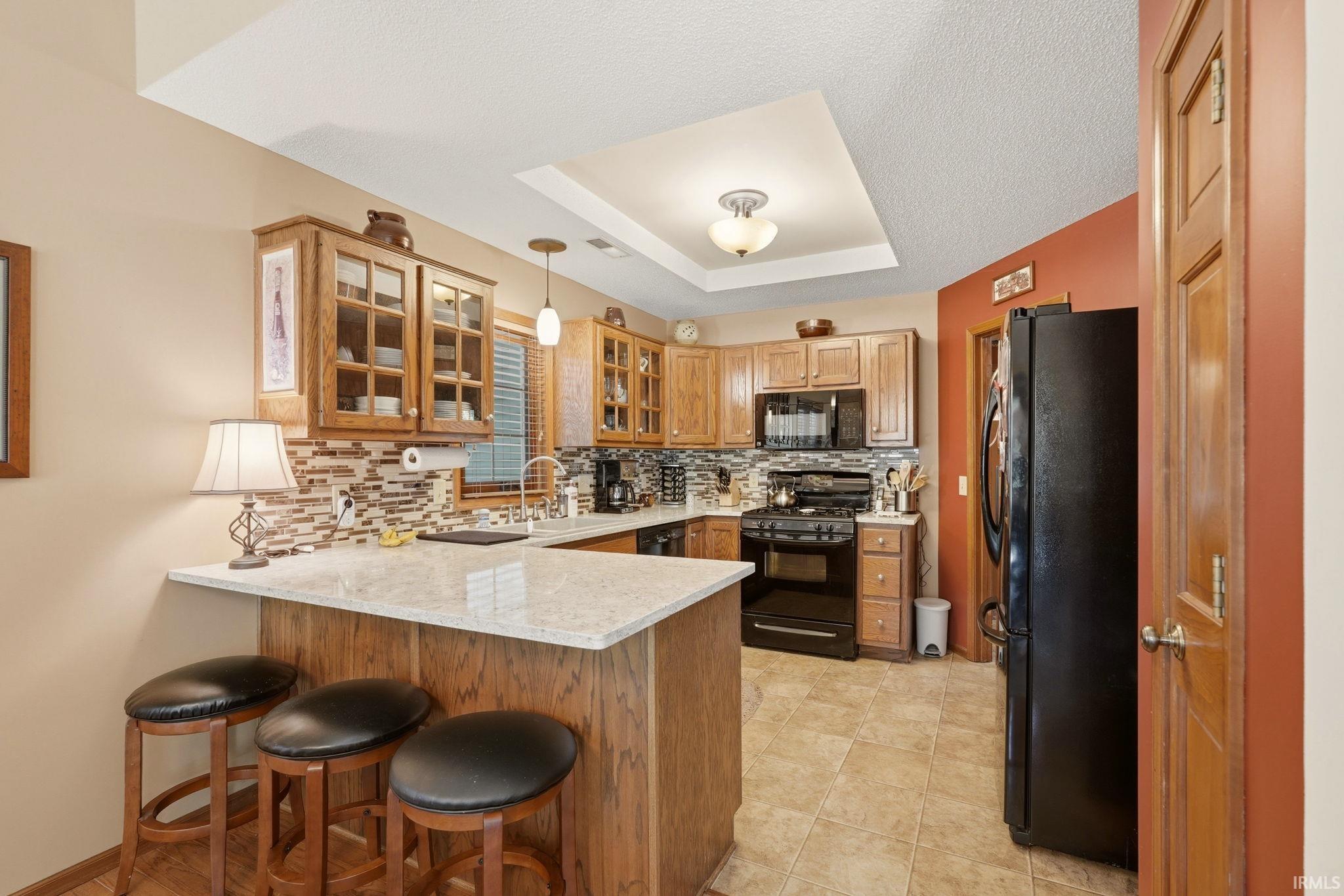 Kitchen featuring a peninsula with a light-toned countertop, wood-finish cabinetry with glass-front uppers, mosaic tile backsplash, black appliances, and a tray ceiling with recessed lighting