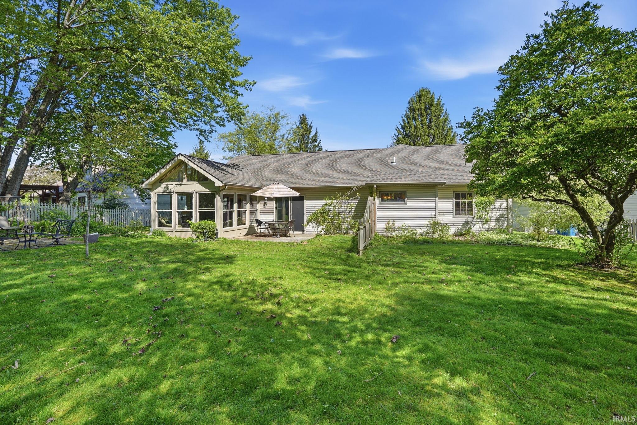 Expansive backyard featuring a manicured lawn, established shade trees, a picket fence, a stone patio, and an enclosed sunroom with multiple windows