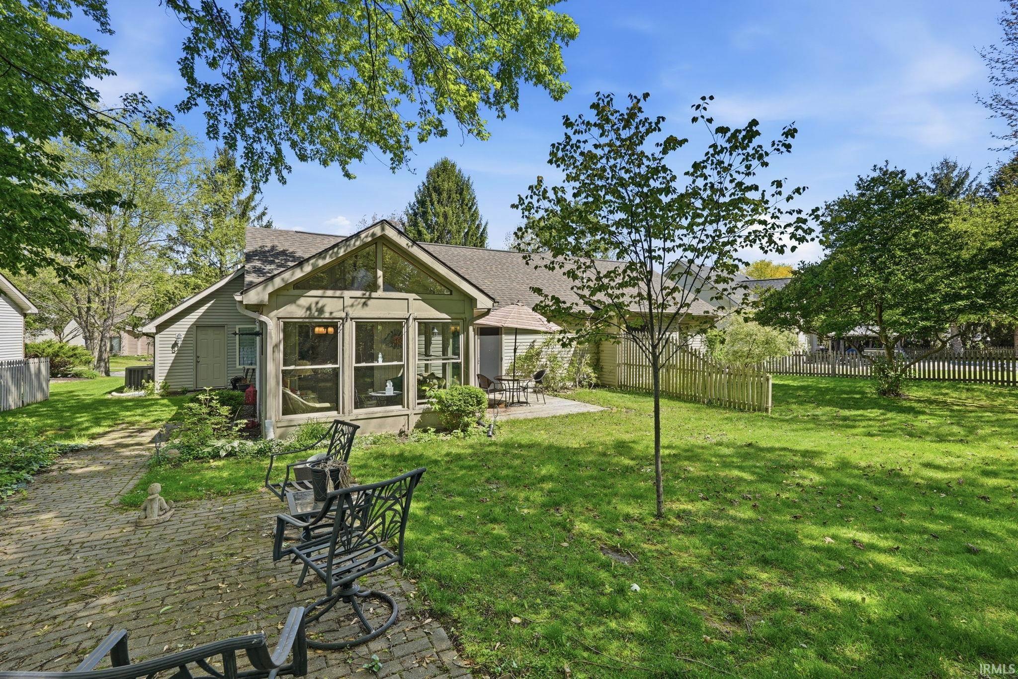 Rear exterior featuring a sunroom with large windows, a paved patio area, a separate concrete patio, a spacious lawn, and mature perimeter trees